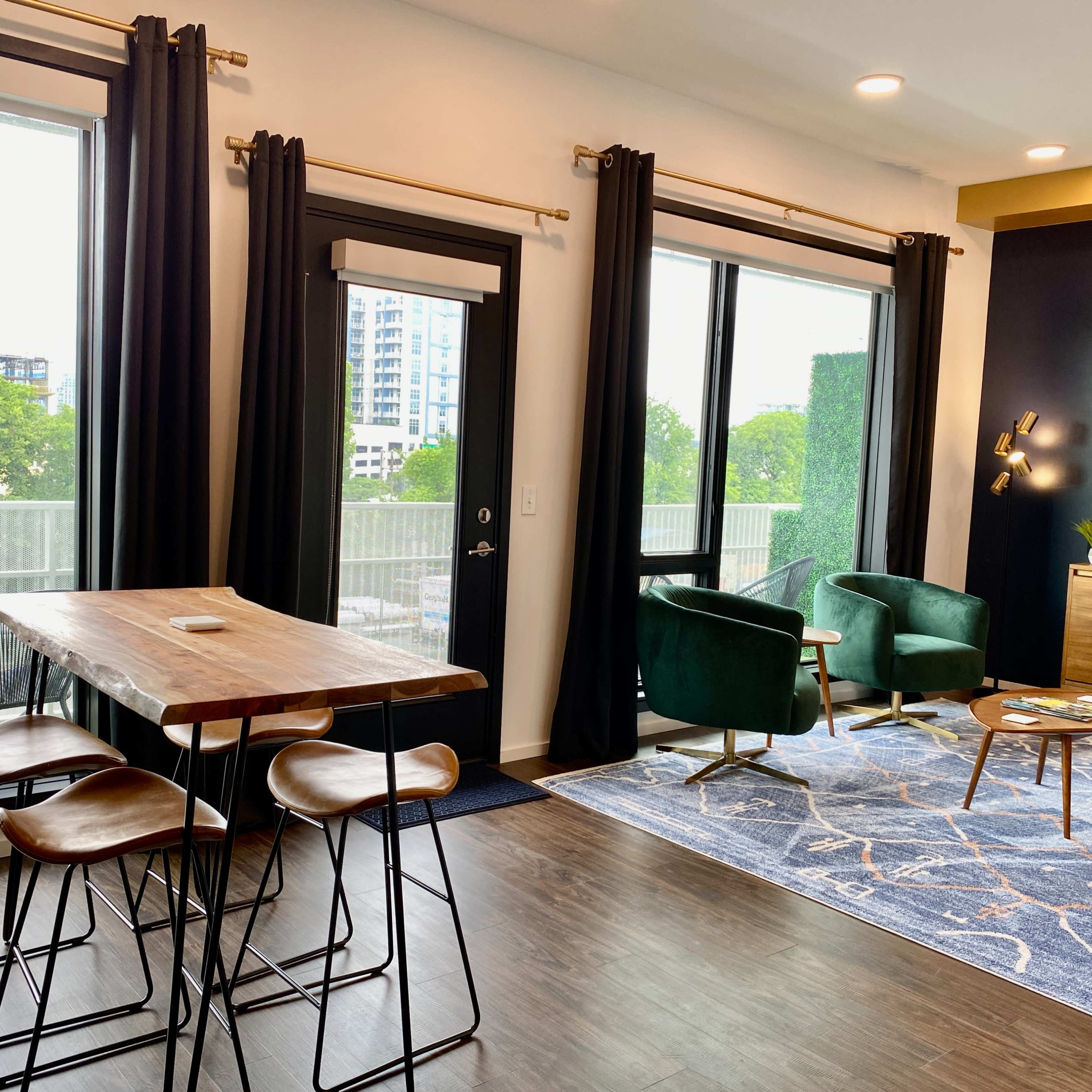A modern apartment living area featuring a wooden dining table with stools, two green accent chairs, a black door leading to a balcony, and large windows letting in natural light.
