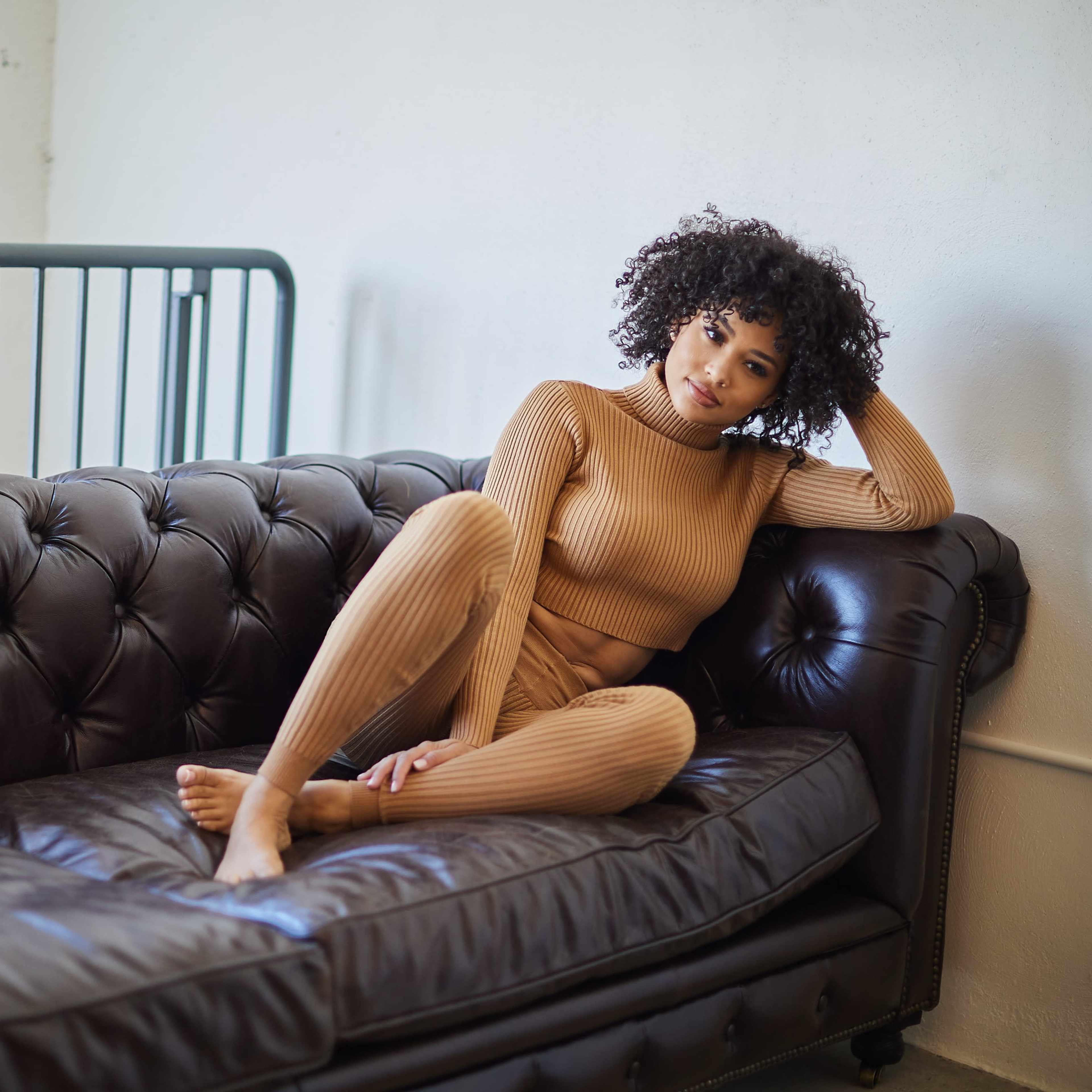 A person in a tan outfit sits casually on a black leather sofa in a well-lit room.