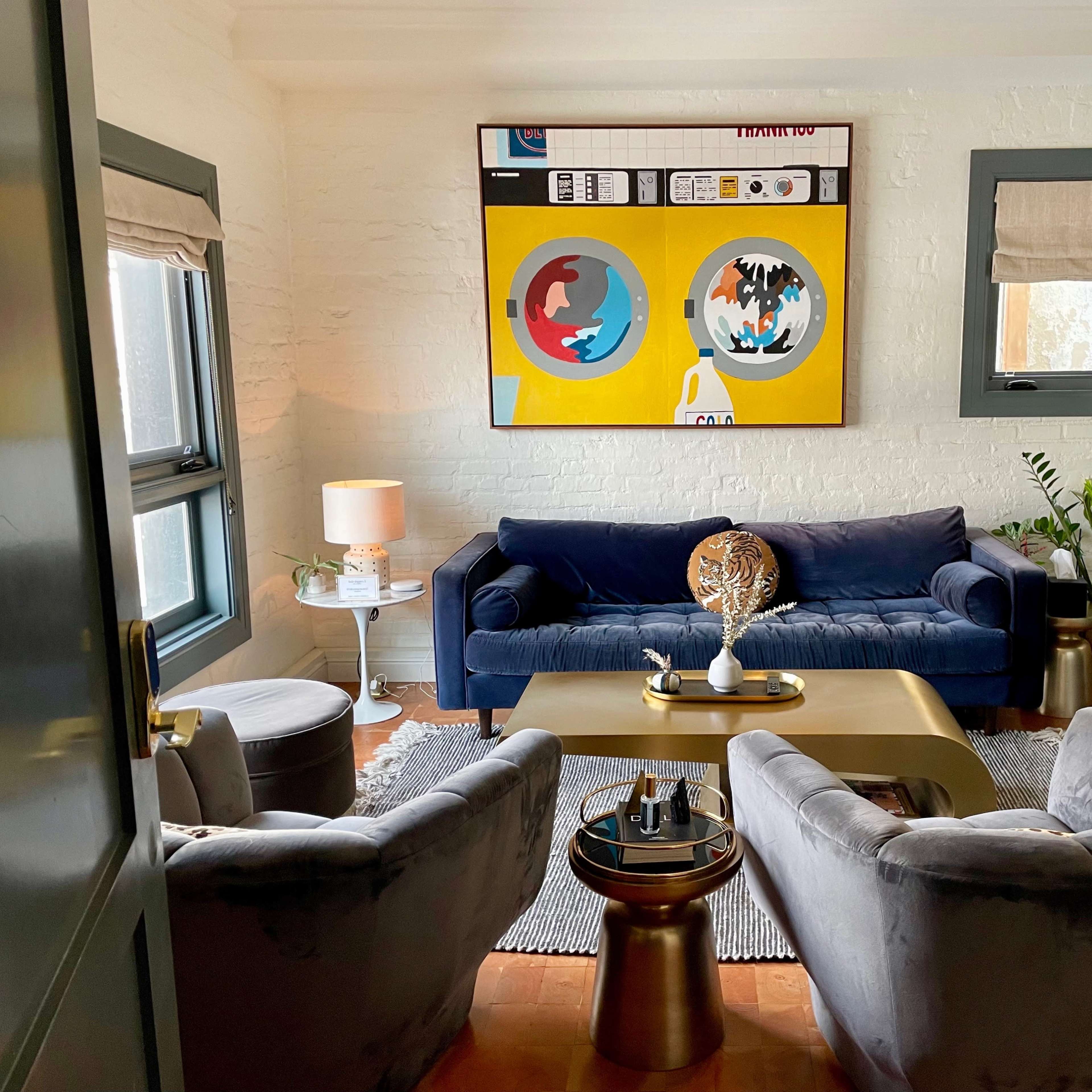 A cozy living room with a blue velvet sofa, two gray armchairs, a gold coffee table, and a colorful wall art piece above the sofa.