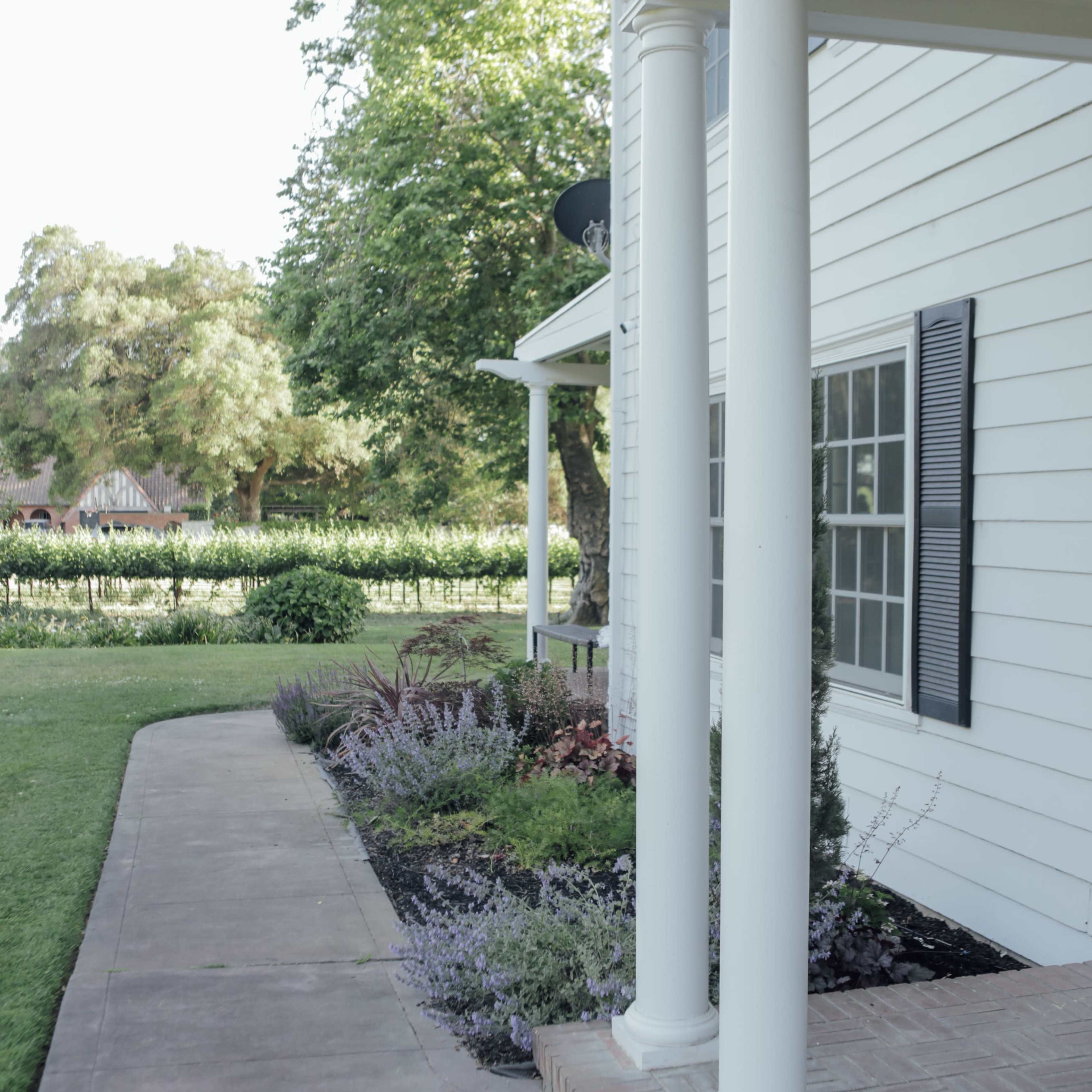 A white house with two columns on a porch, surrounded by a landscaped garden along a concrete pathway.
