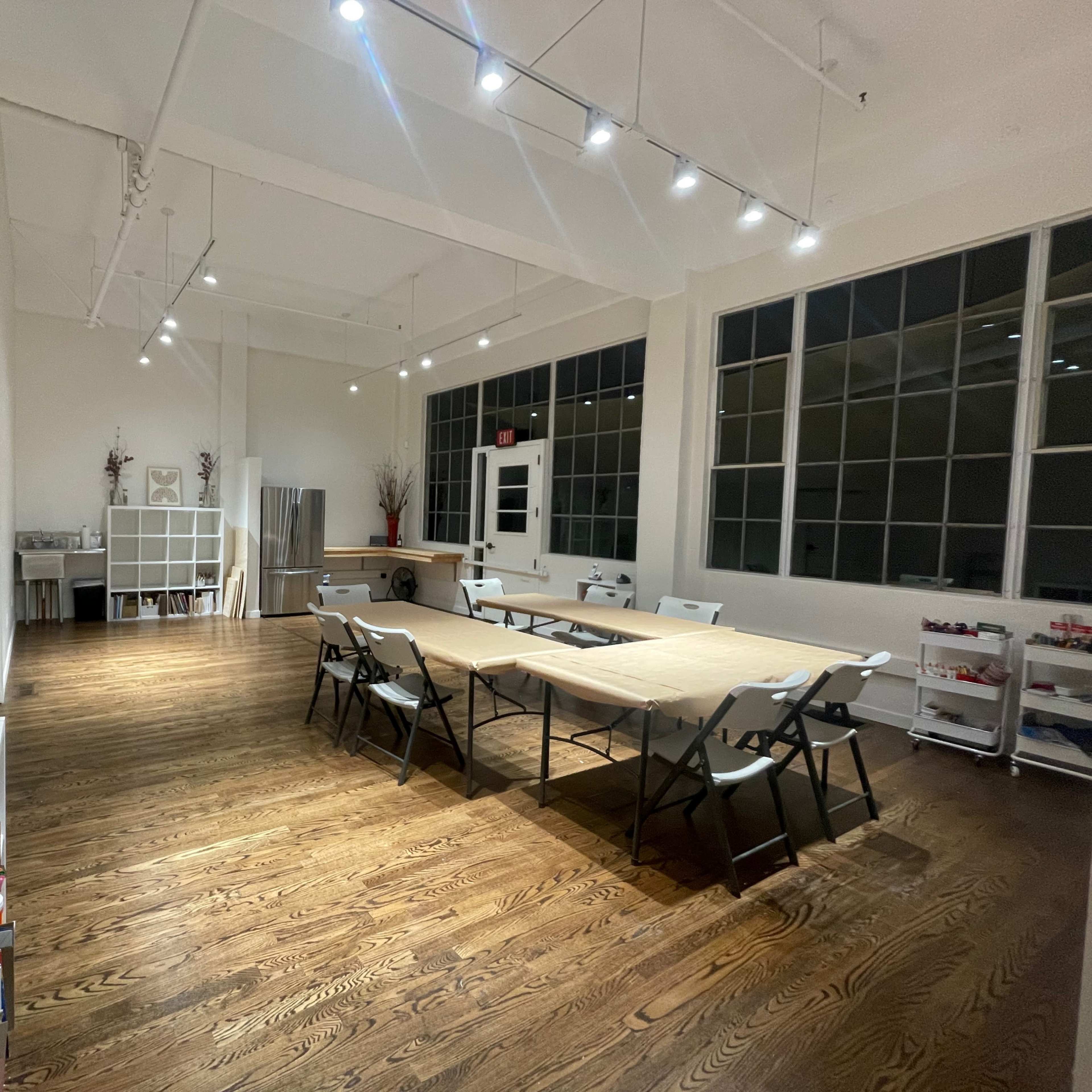 A spacious room with wooden floors, large windows, and a central table surrounded by folding chairs, with shelves and a decorative vase along the walls.