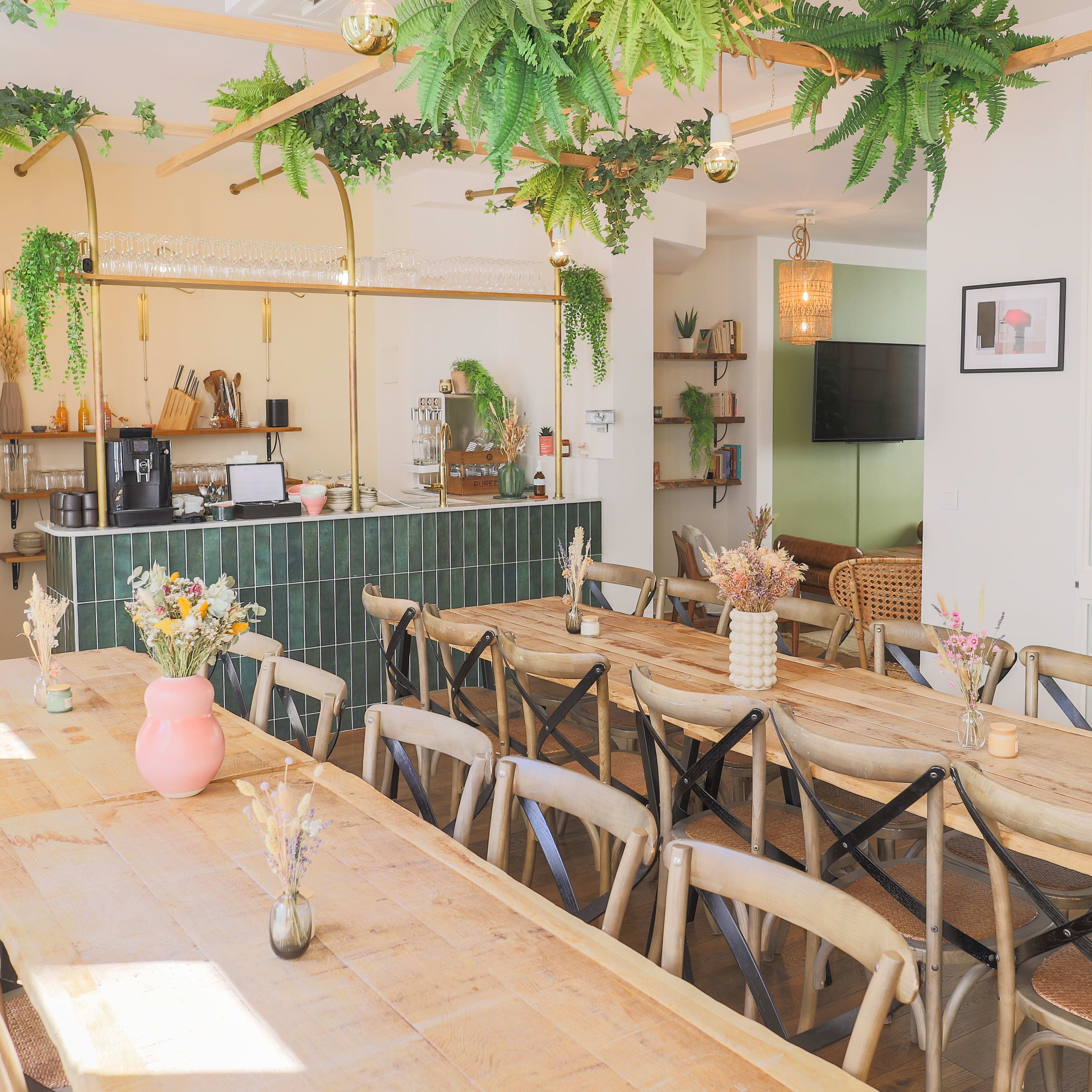 A spacious, well-lit cafe features a long wooden table surrounded by chairs, with greenery hanging from the ceiling and a counter in the background.