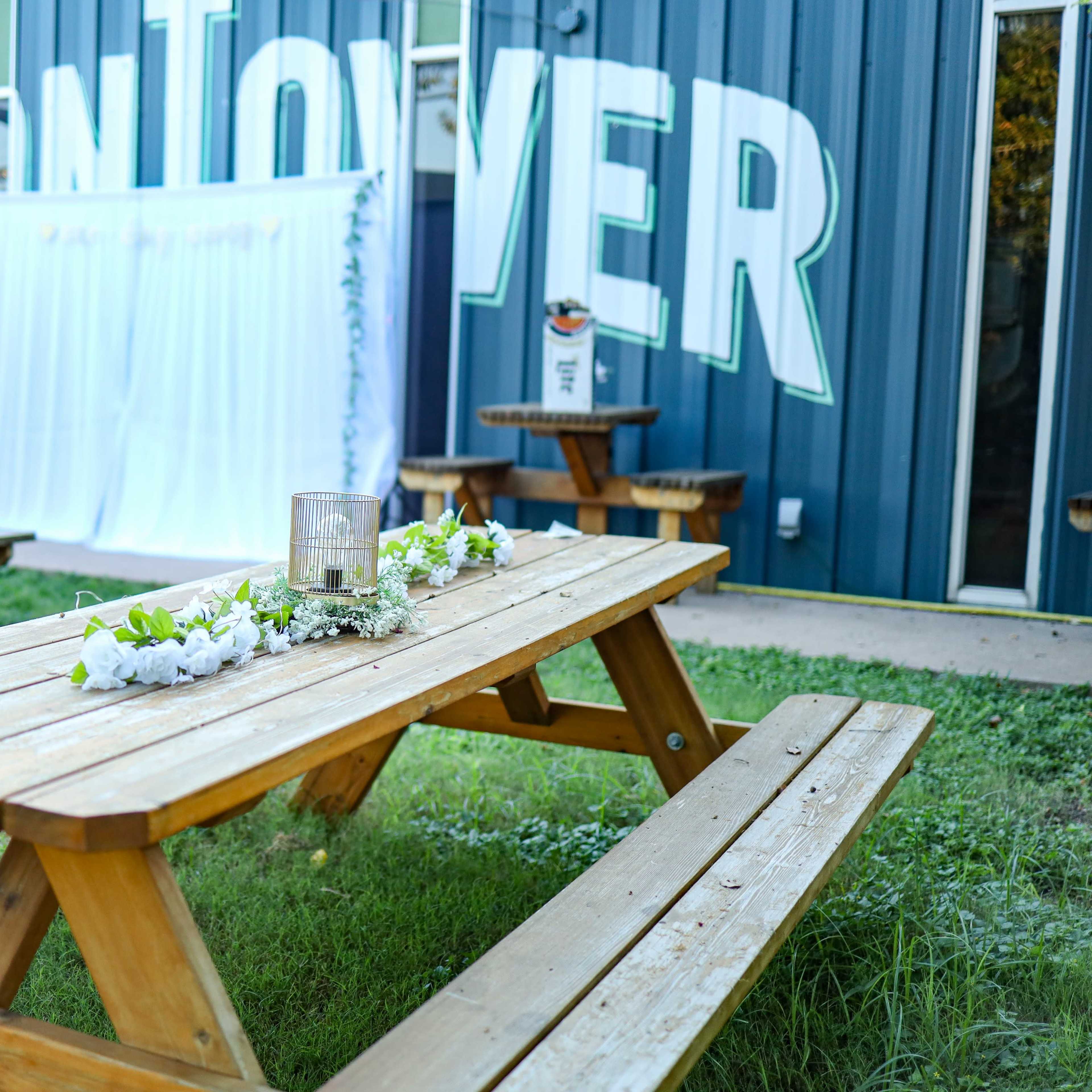 A set of wooden picnic tables with floral arrangements is placed in front of a building featuring large letters that read "MONTOVER."