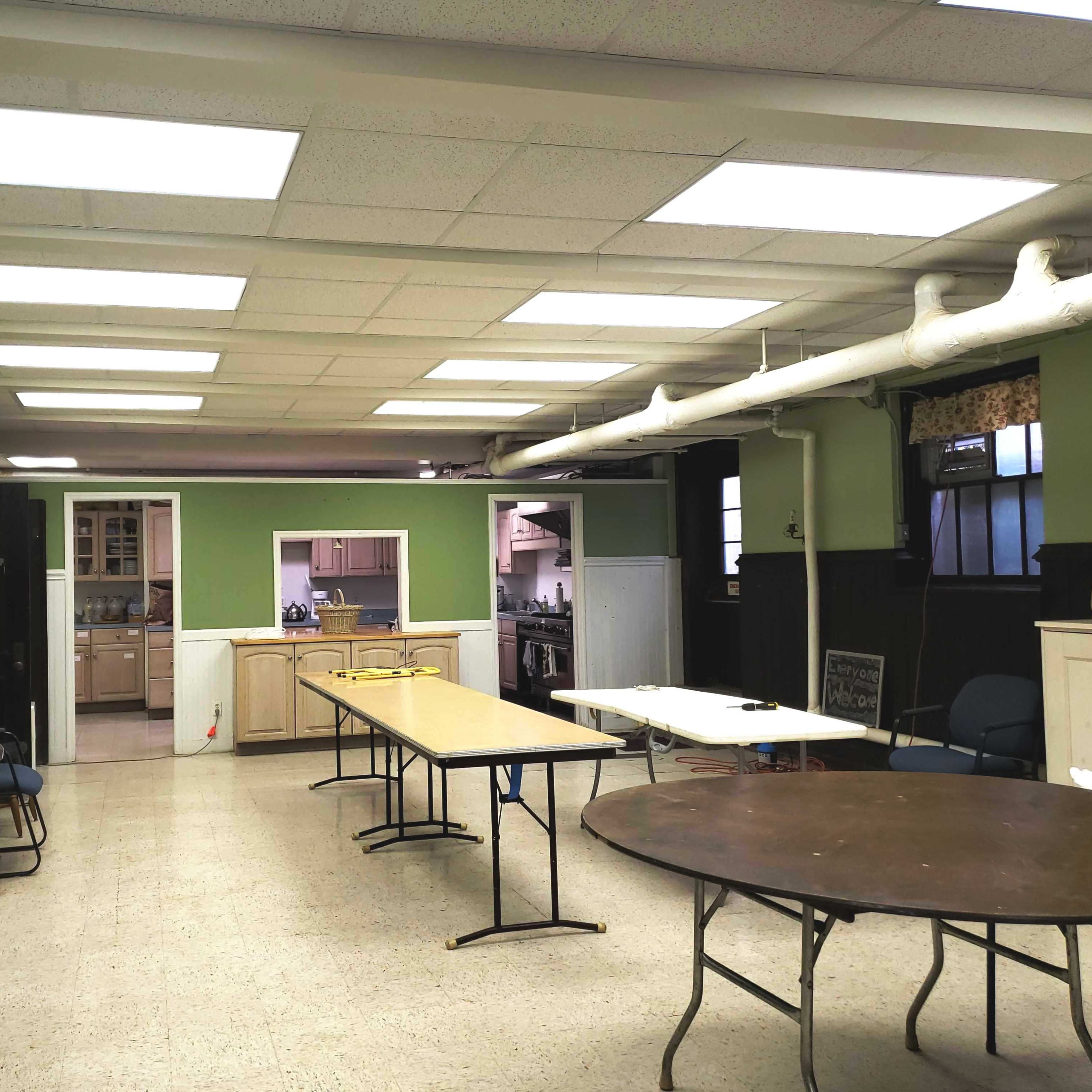An empty room with several tables arranged, light-colored walls, and a kitchen area visible in the background.
