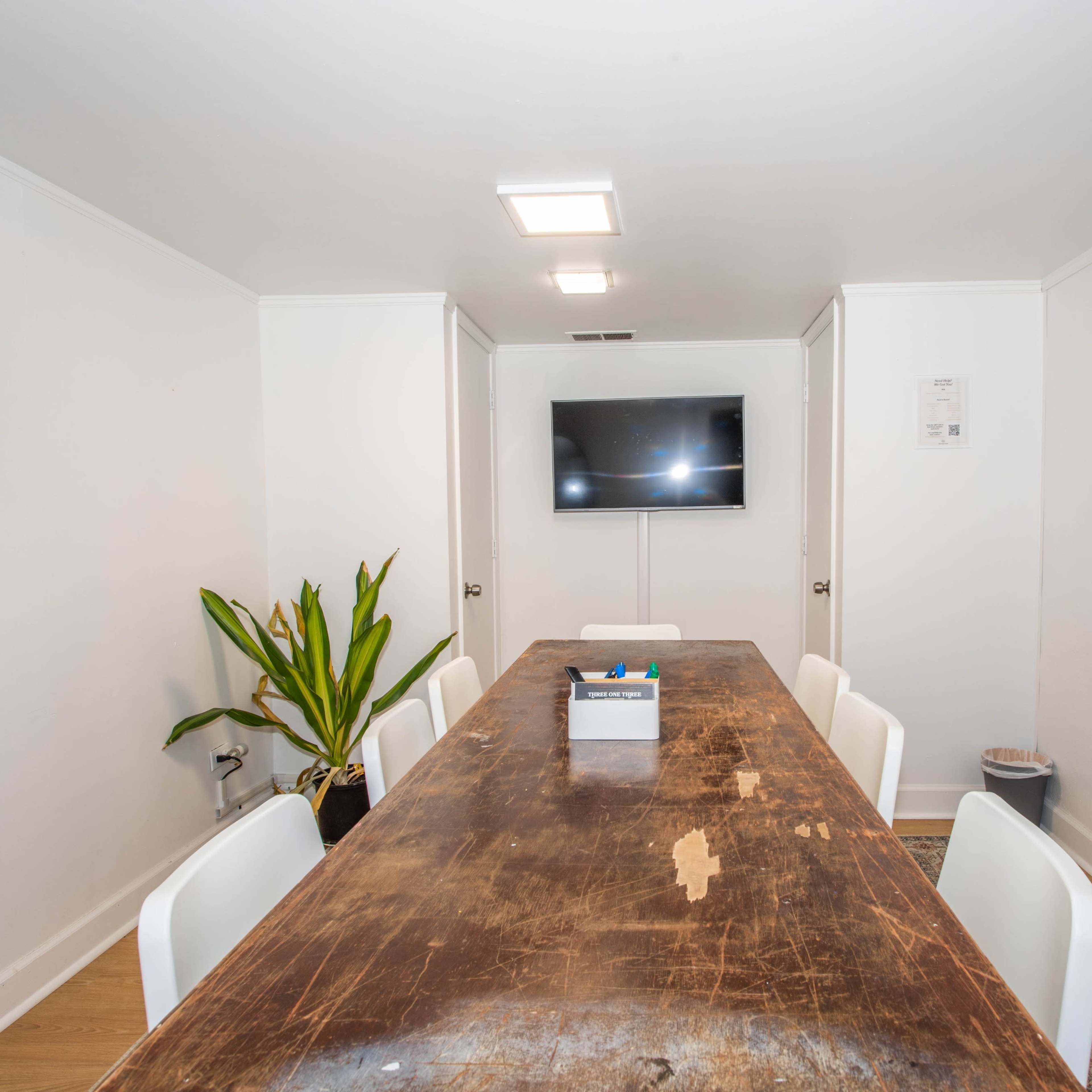 A long, wooden table is surrounded by white chairs in a well-lit meeting room featuring a television and a whiteboard on the walls.