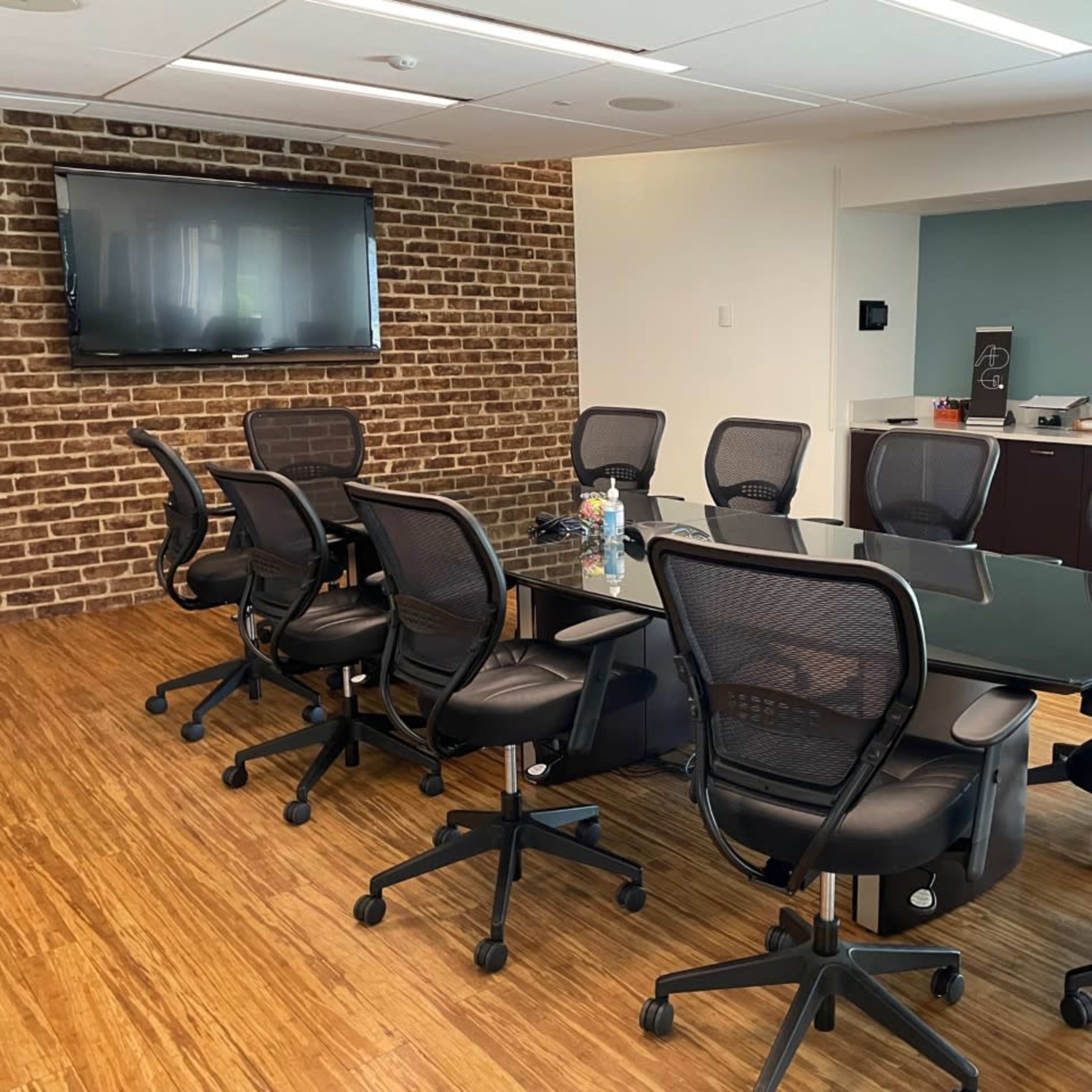 A conference room features a large glass table surrounded by black mesh chairs, with a brick wall and a television mounted on one side.