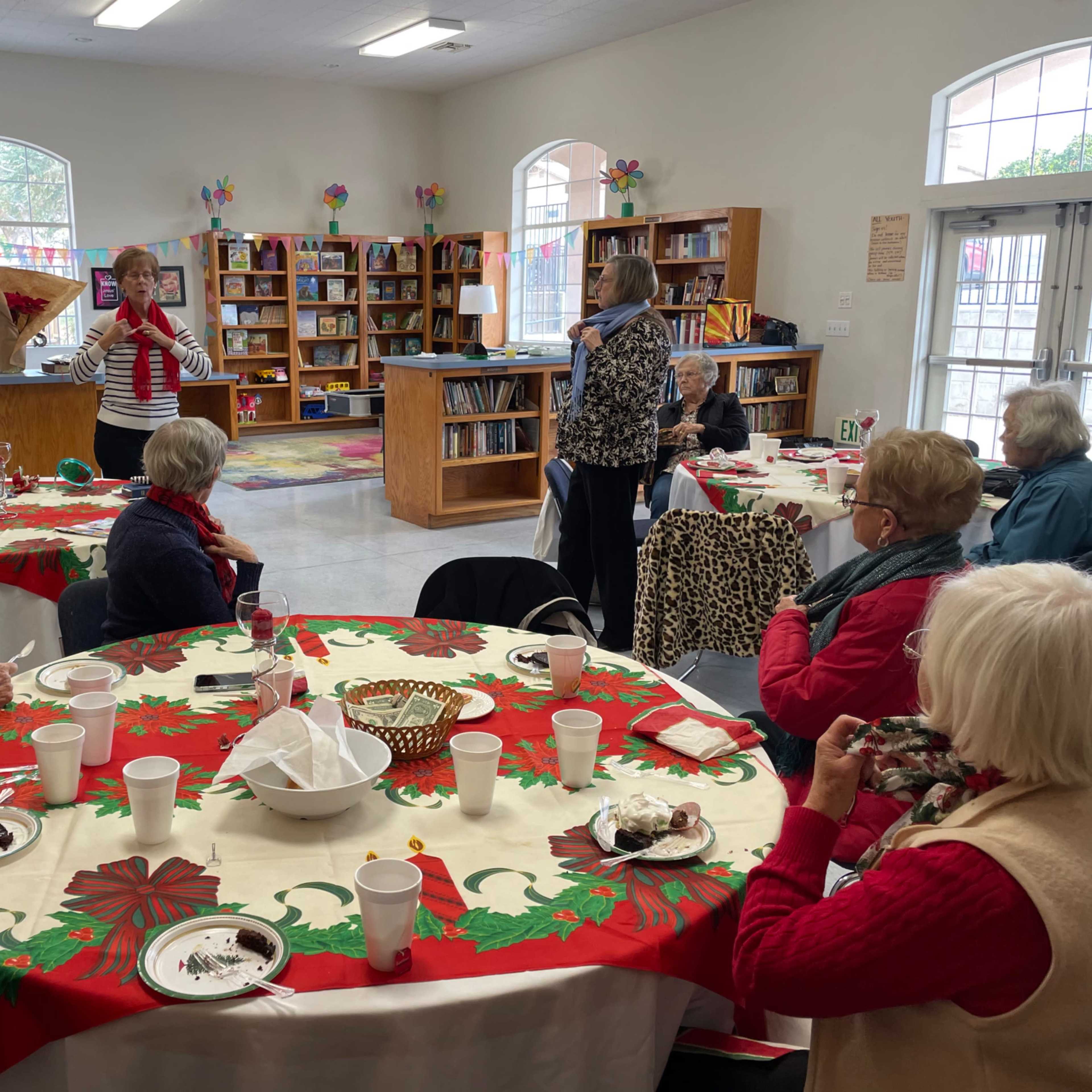A group of seniors gathers around tables decorated for a festive event in a community room with bookshelves and colorful decorations.