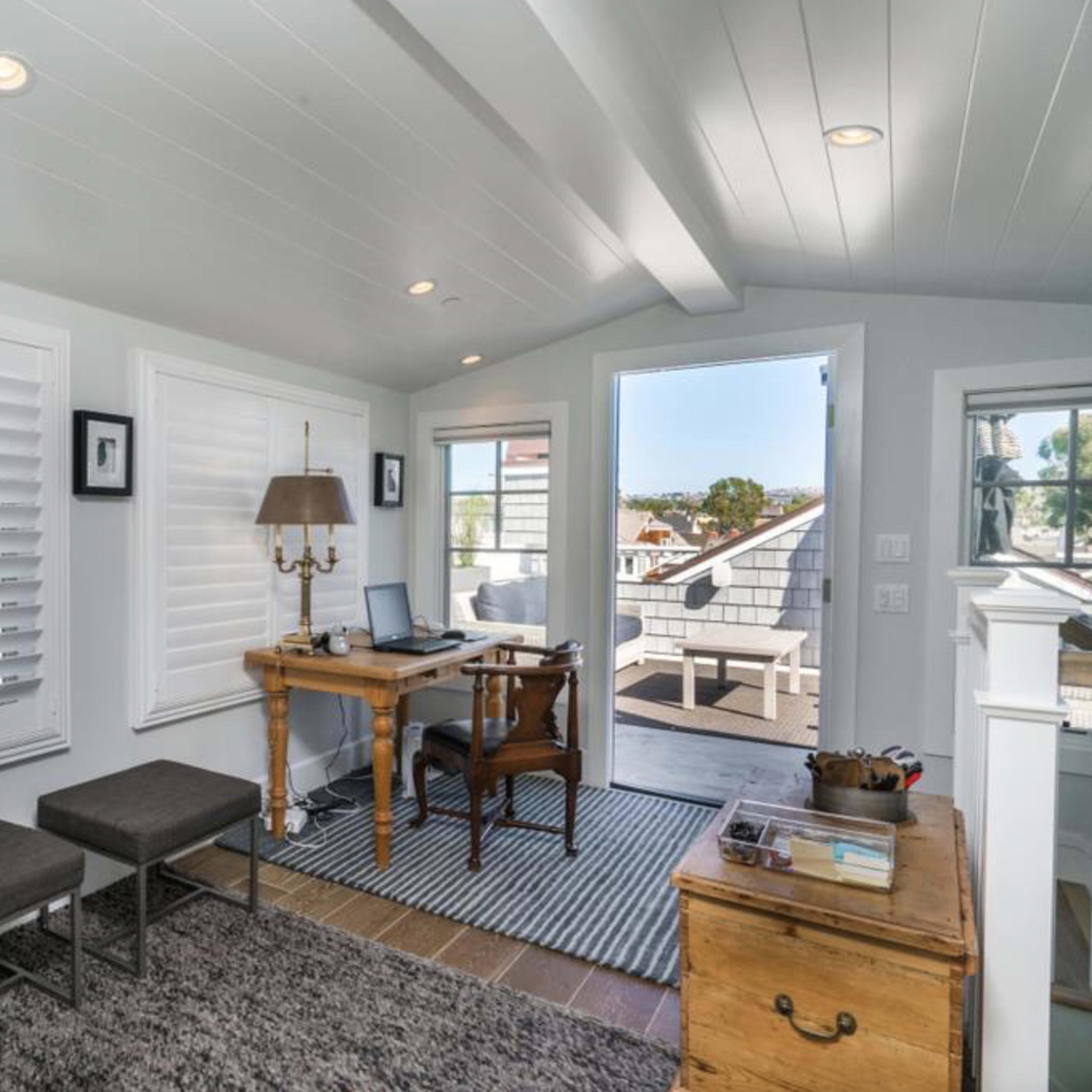 A bright home office features a wooden desk, two benches, and large windows with shutters, opening to a small balcony.