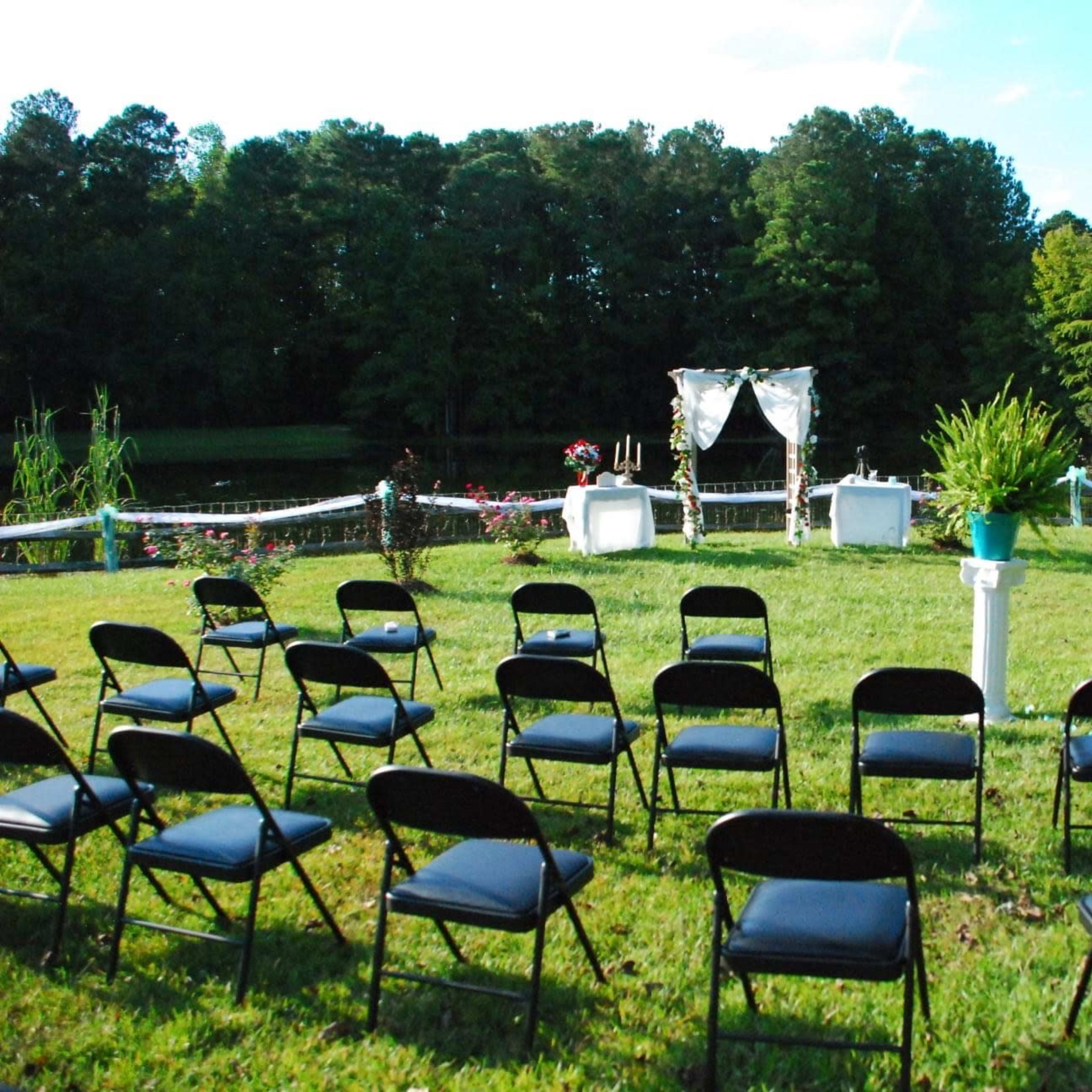 A grassy area set up for a wedding ceremony, featuring rows of black chairs facing a decorated altar with a backdrop of trees and a body of water.
