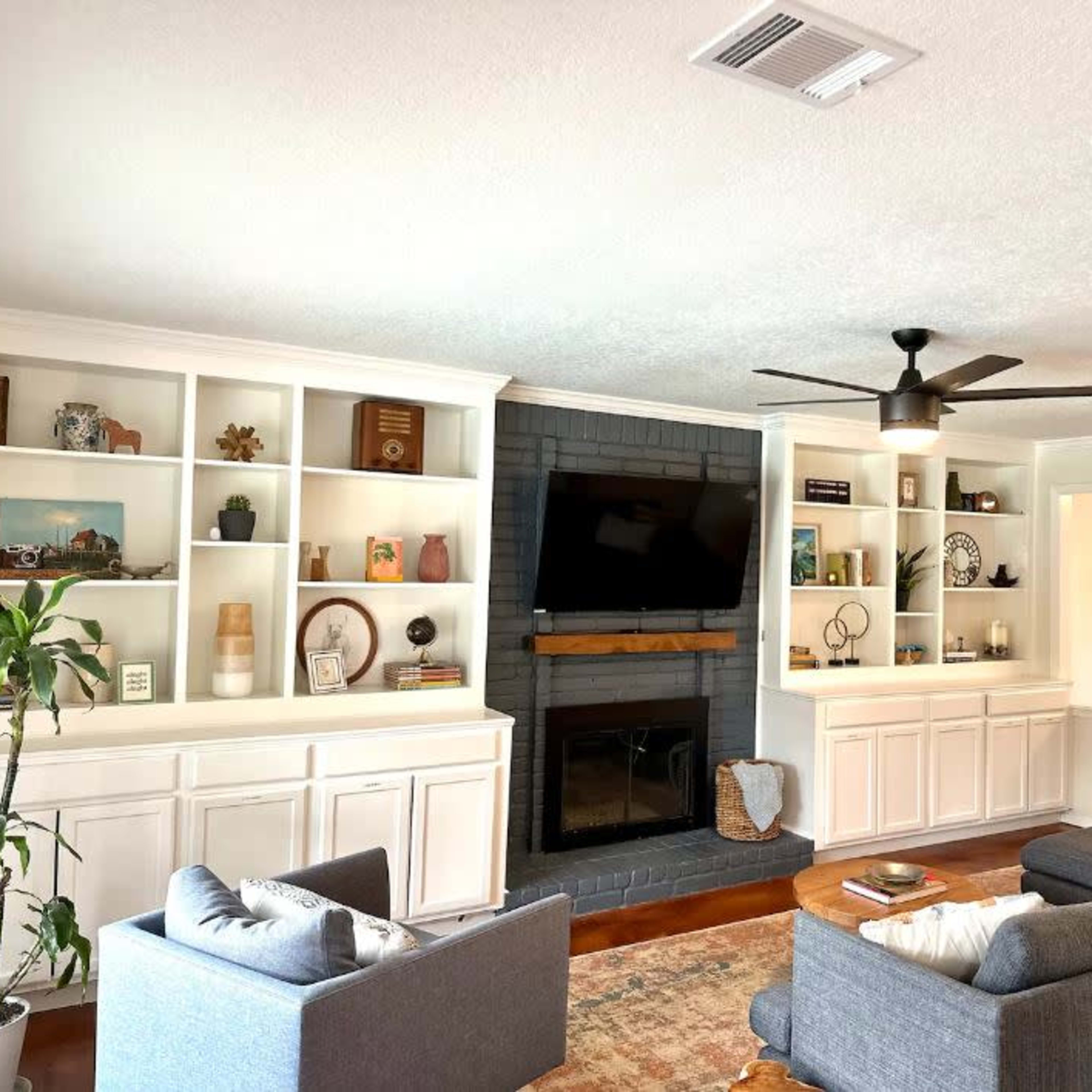 A living room featuring two gray sofas, a dark brick fireplace, built-in shelving with decorative items, and a ceiling fan.