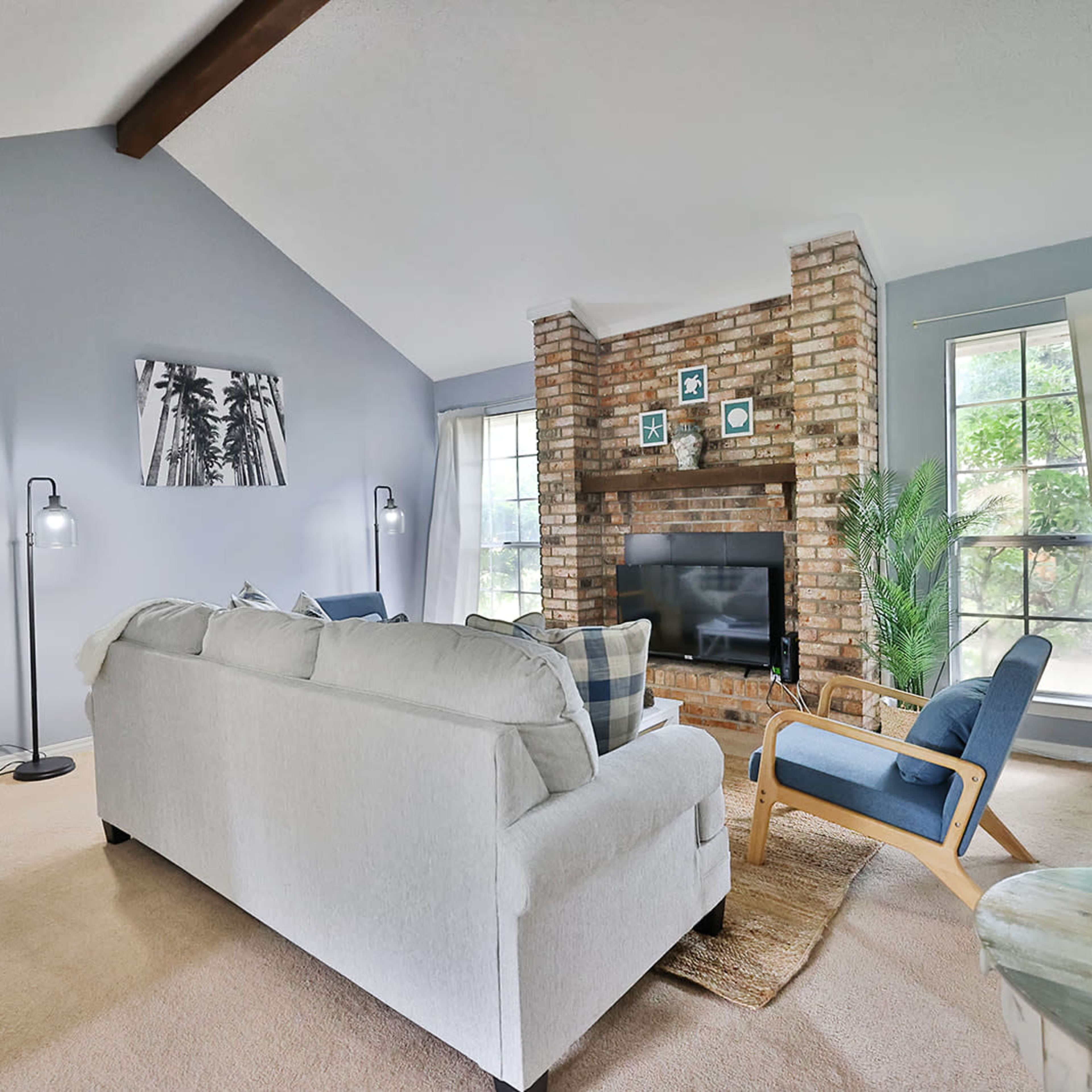 A living room featuring a white sofa, blue accent chair, and a brick fireplace, with large windows and light-colored walls.