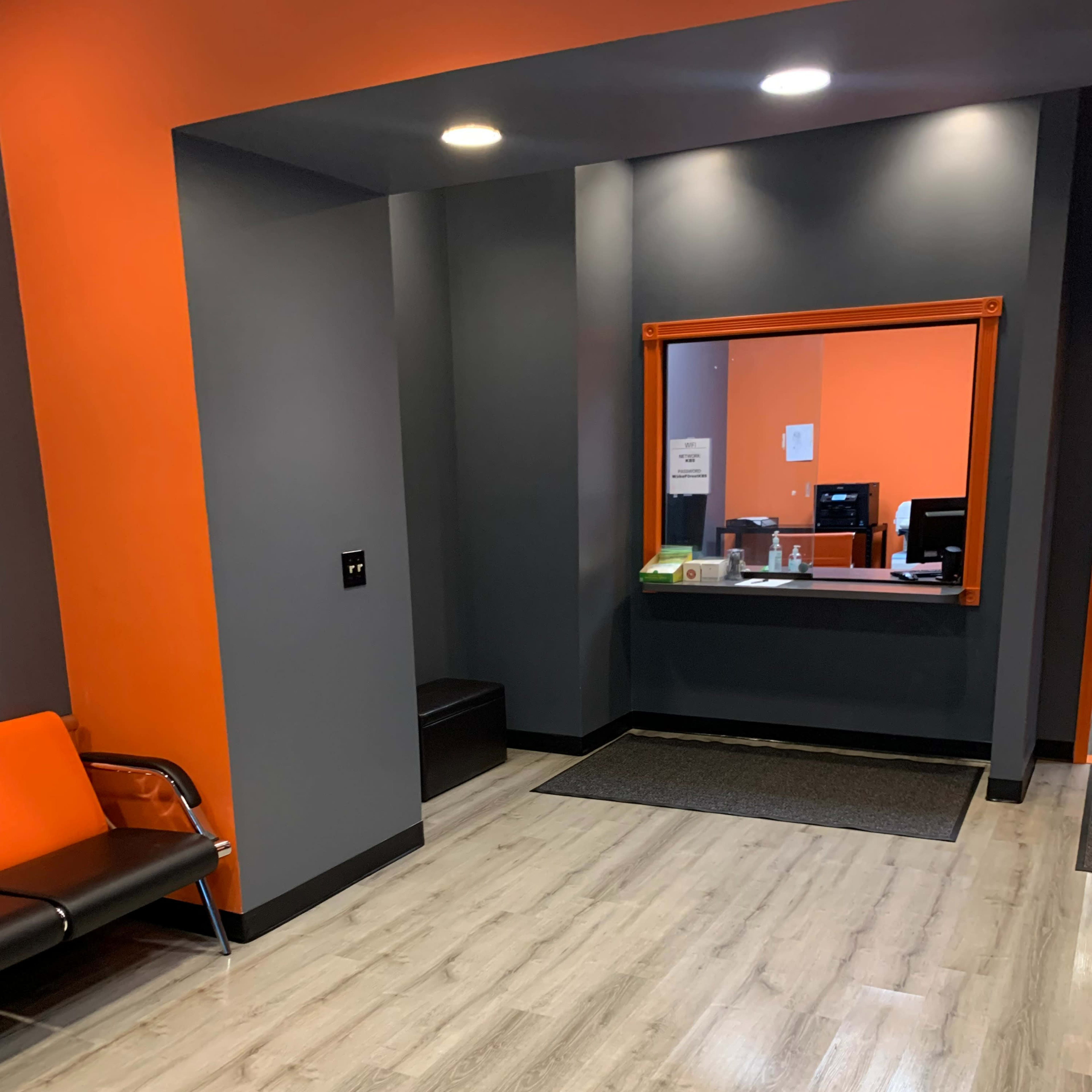 A modern reception area with gray walls, orange accents, a seating area, and a window counter leading to a workspace.