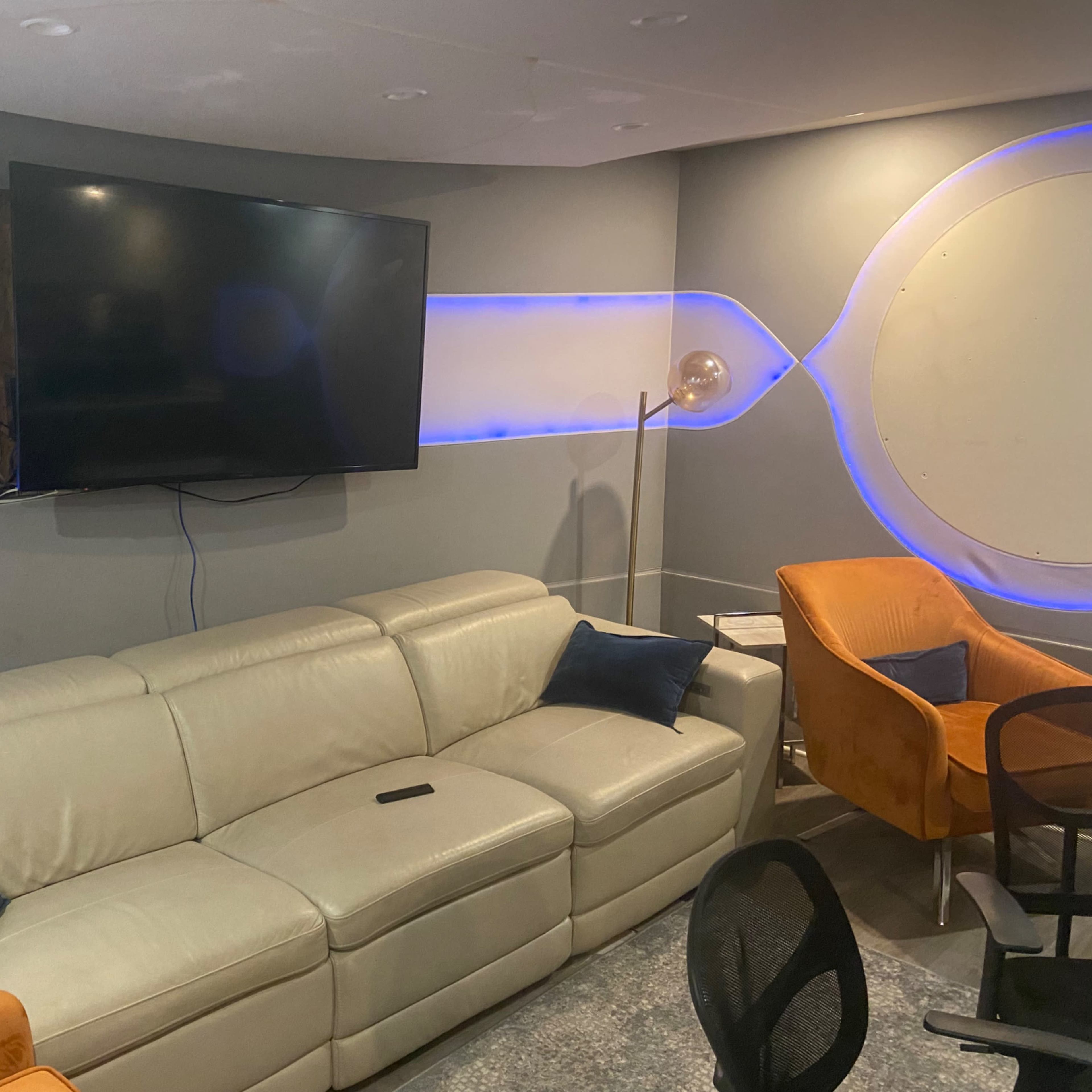 A modern, minimalist living room with a sectional beige sofa, a large flat-screen TV mounted on the wall, and two orange armchairs.
