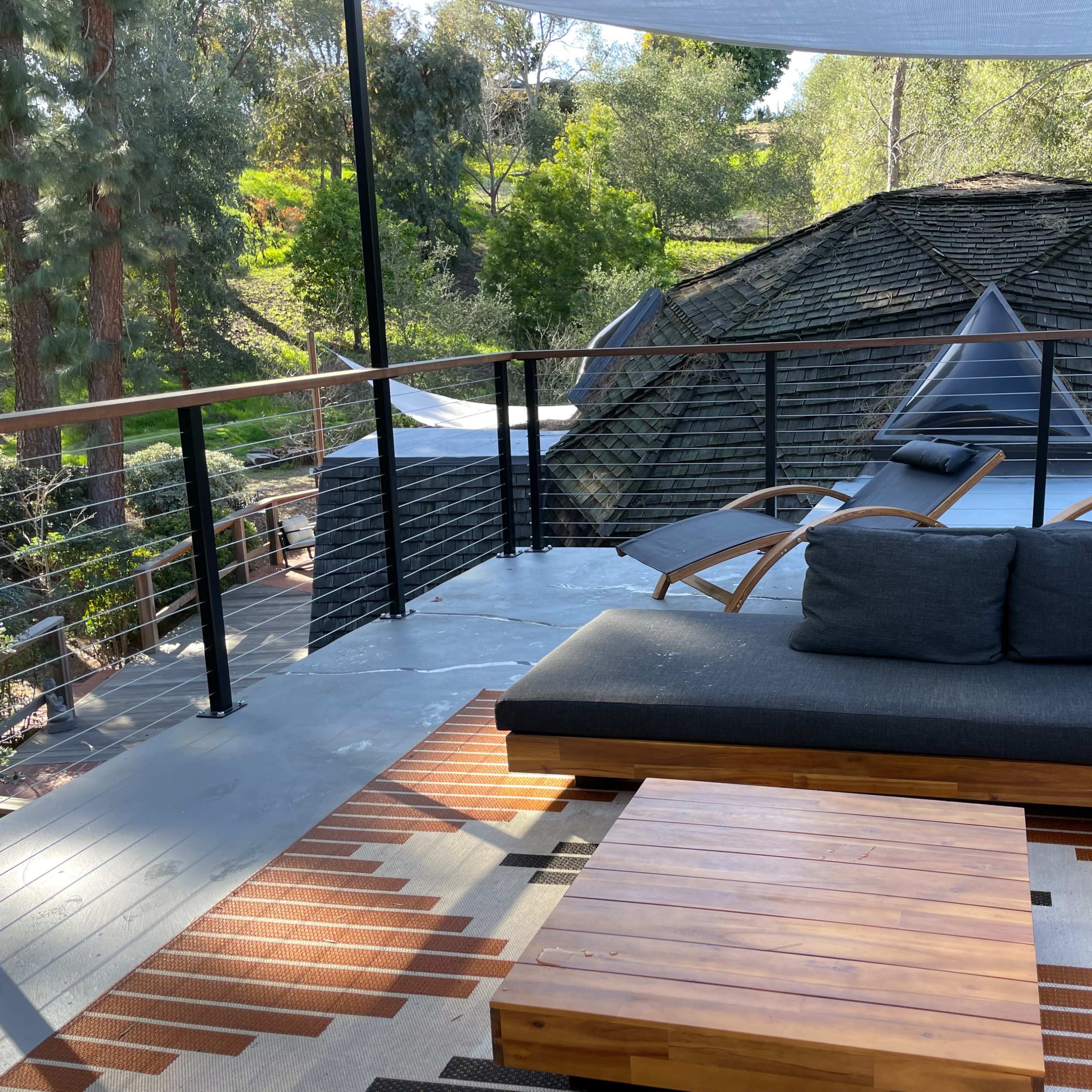 A wooden deck features a sectional sofa, a coffee table, and two lounge chairs overlooking a wooded area and a distinctive roof structure.
