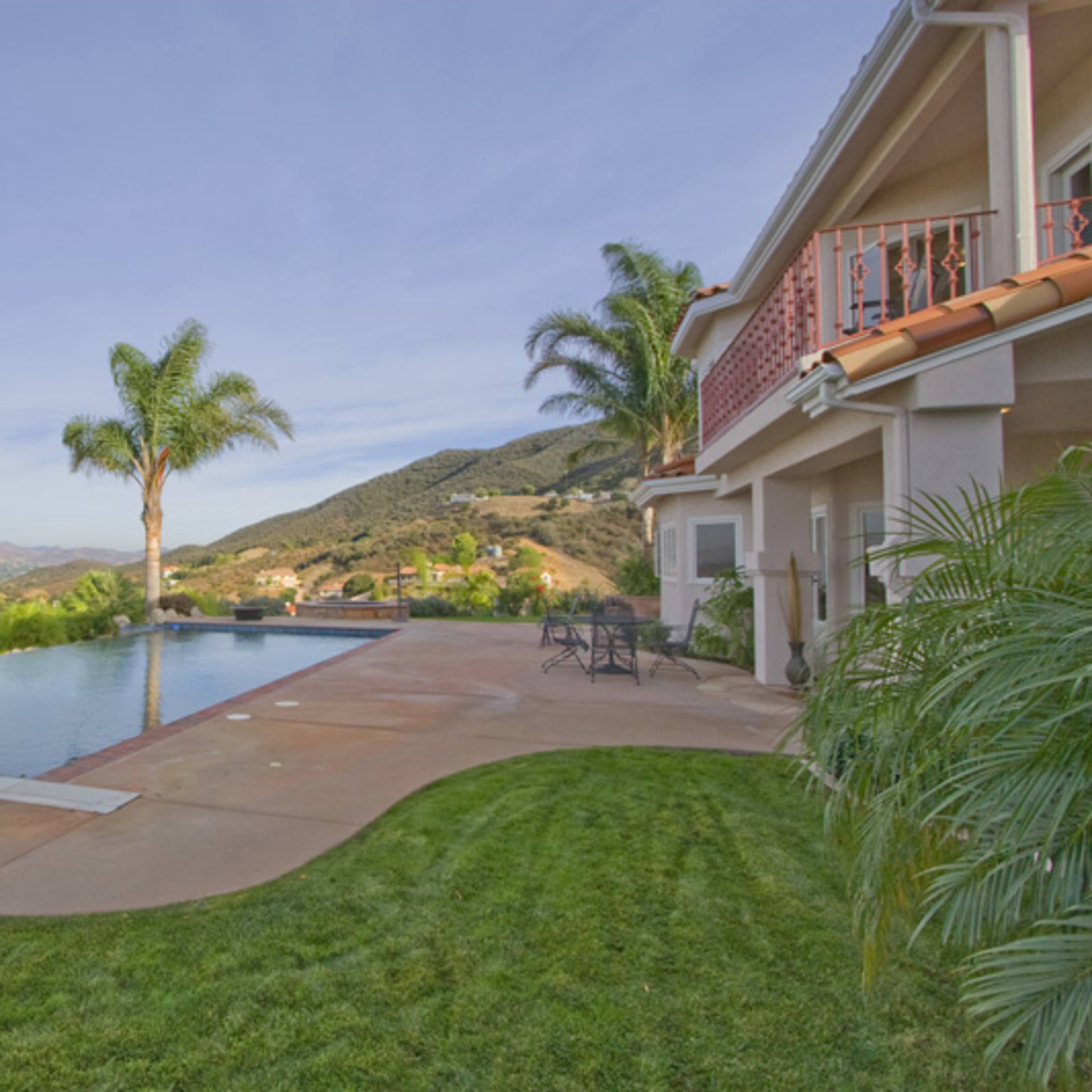 A modern house sits beside an infinity pool overlooking a mountainous landscape with palm trees and a lawn.