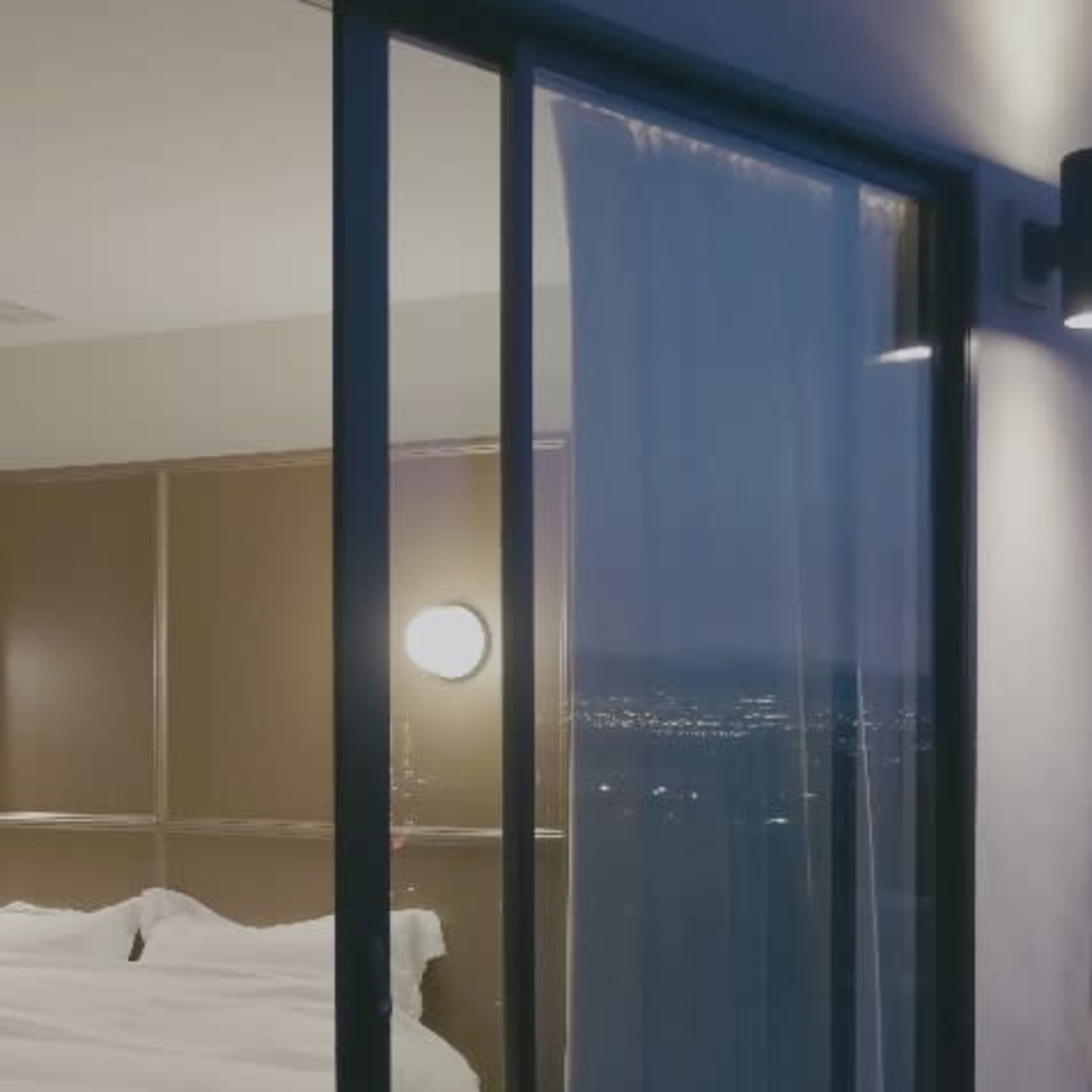 A balcony door opens to a bedroom with a neatly made bed and a city view at dusk.
