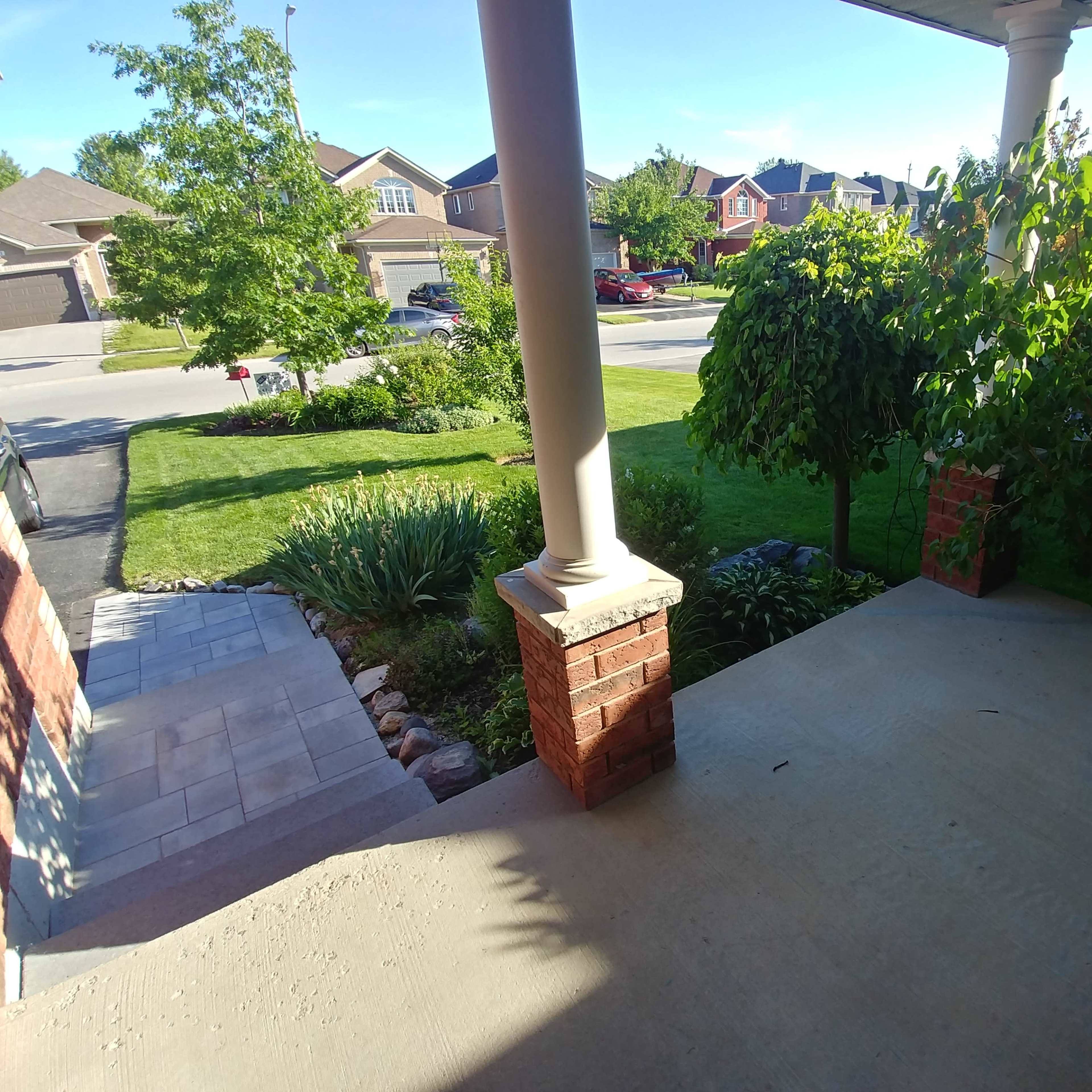 A front porch view with a concrete floor, brick columns, and landscaping featuring grass, shrubs, and a tree.
