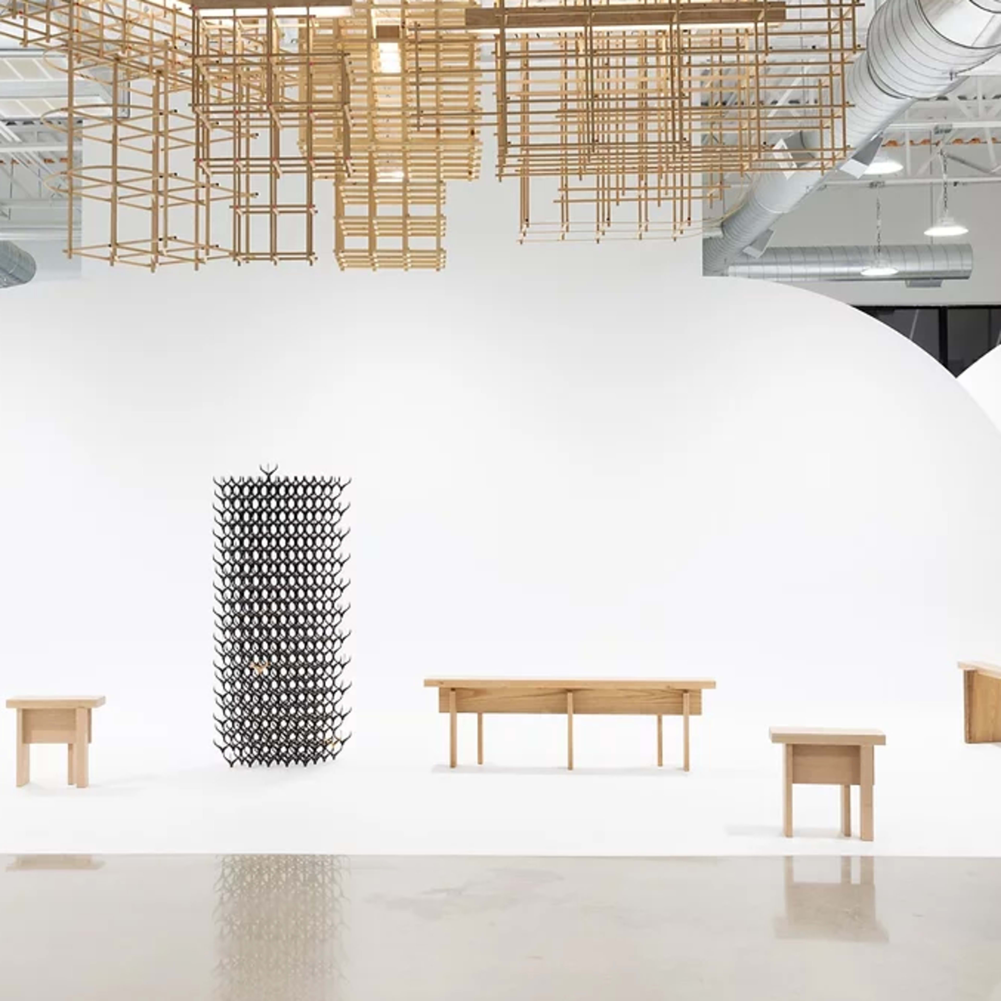 A minimalist art space featuring a central sculpture constructed from a grid of black shapes, surrounded by wooden tables against a white backdrop.