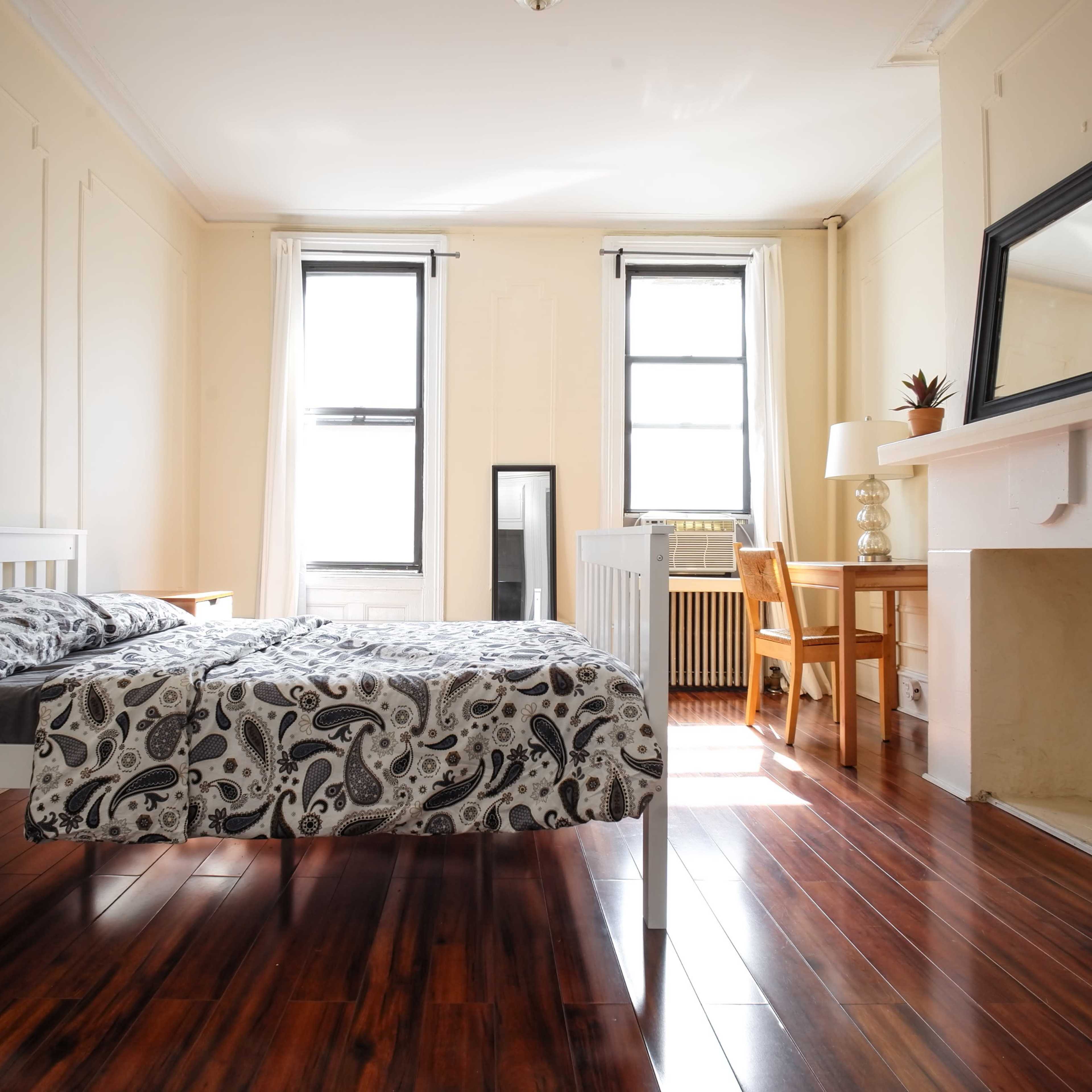 The room features a bed with a patterned comforter, a wooden chair and table, a fireplace, and large windows allowing natural light to enter.