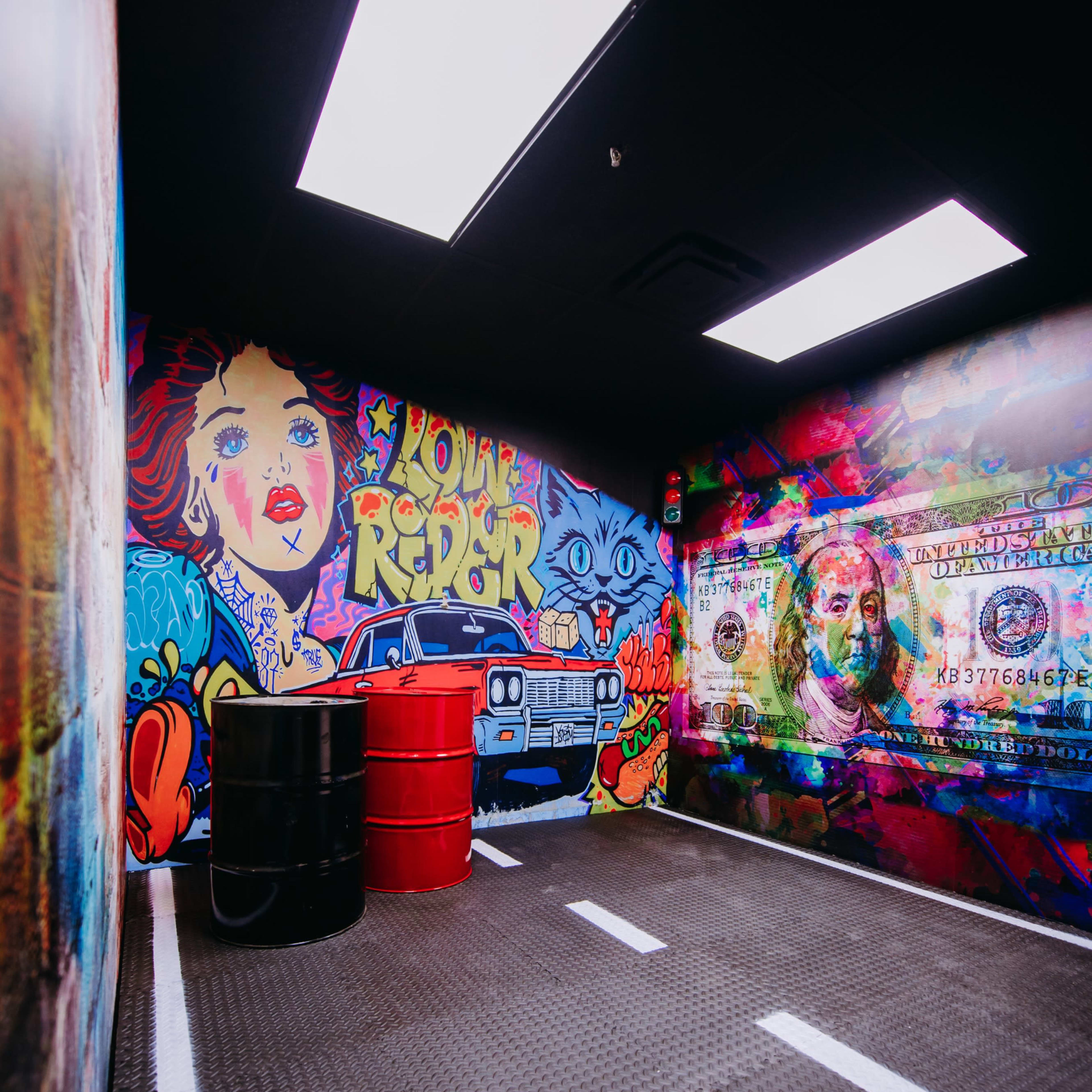 A dimly lit room with vibrant graffiti on the walls, featuring a vintage car design and two red barrels.