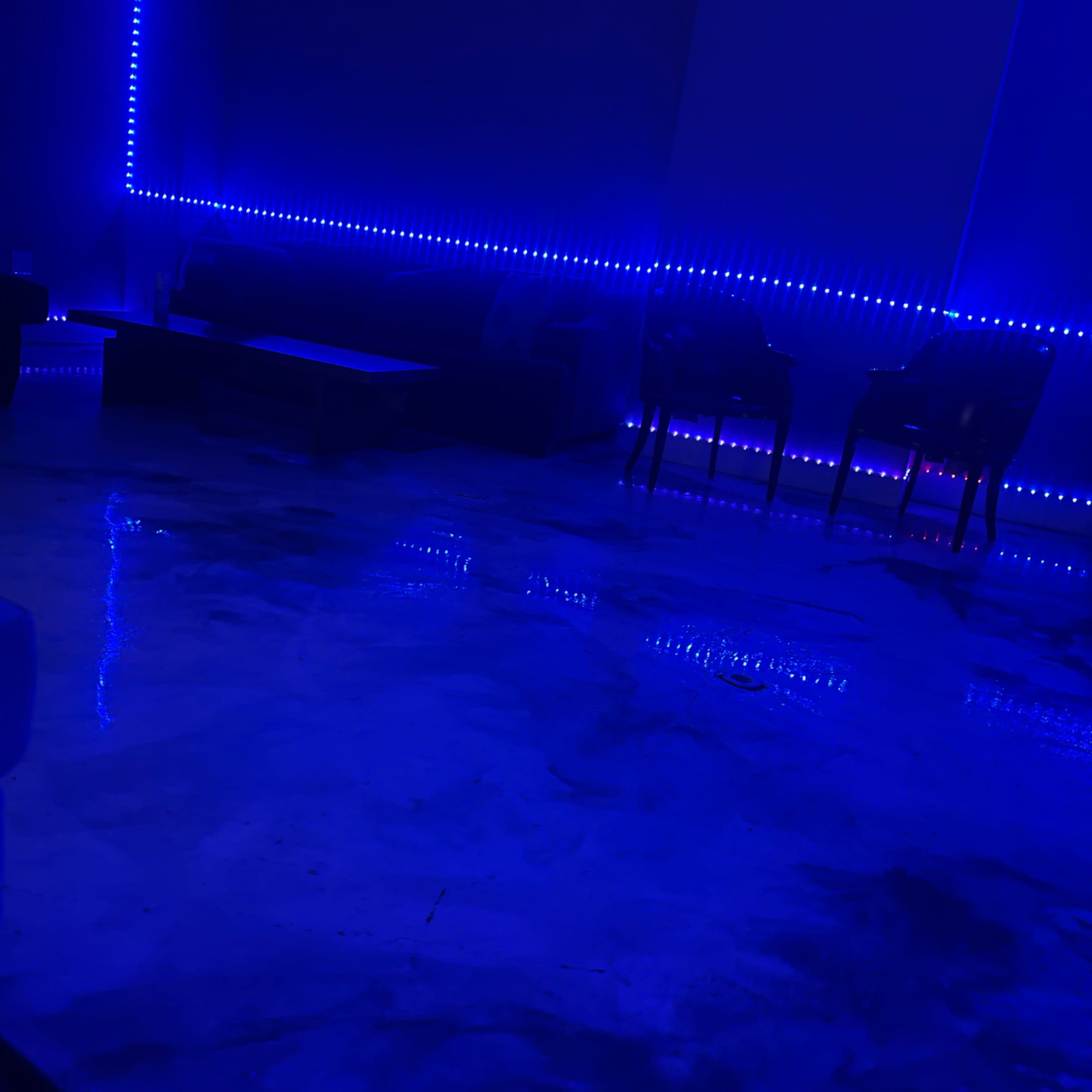 A dimly lit room features blue LED lights illuminating a smooth, reflective floor with a sofa and two chairs along the walls.
