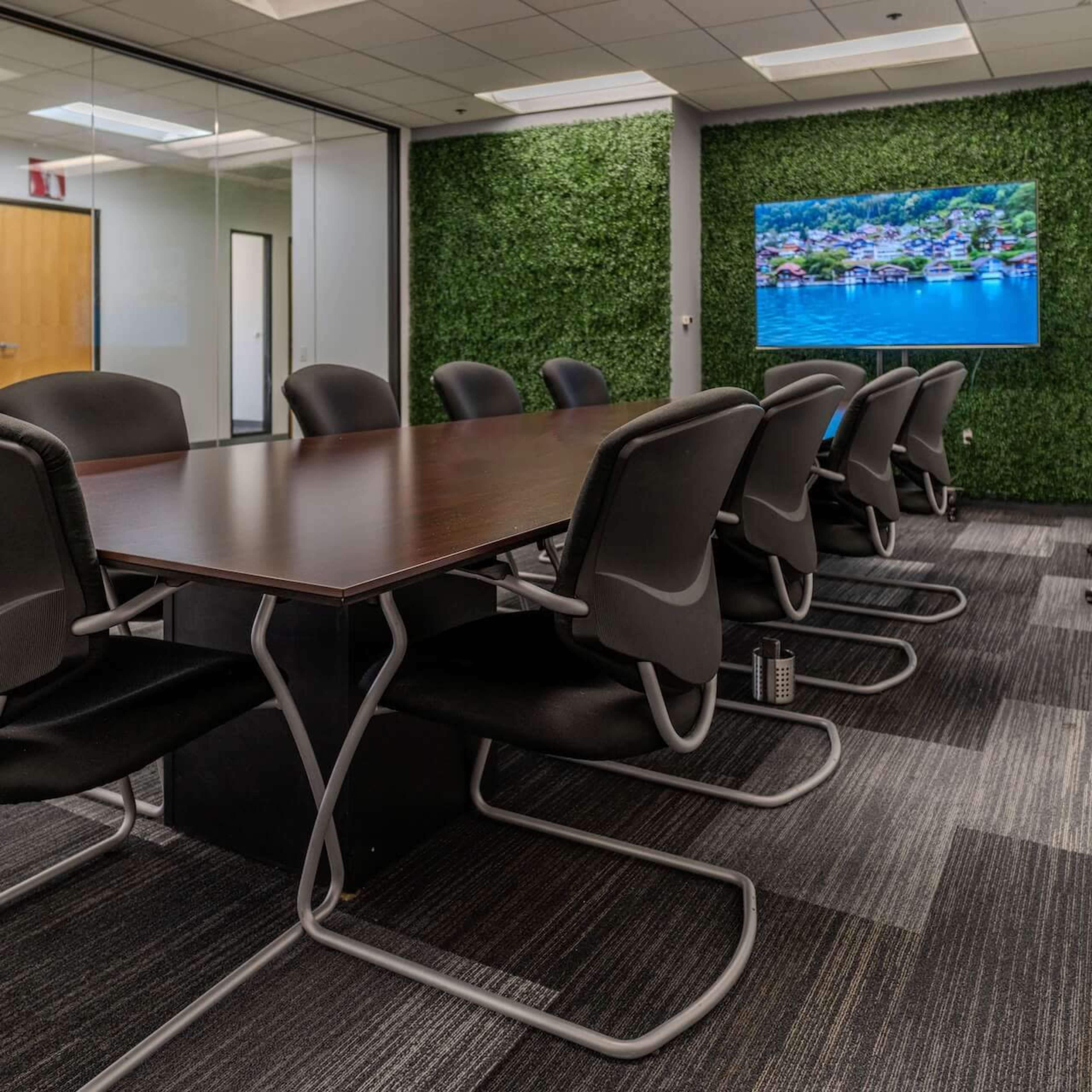 A modern conference room features a long wooden table surrounded by black chairs, with glass walls and a large screen displaying a scenic image of a coastal village.