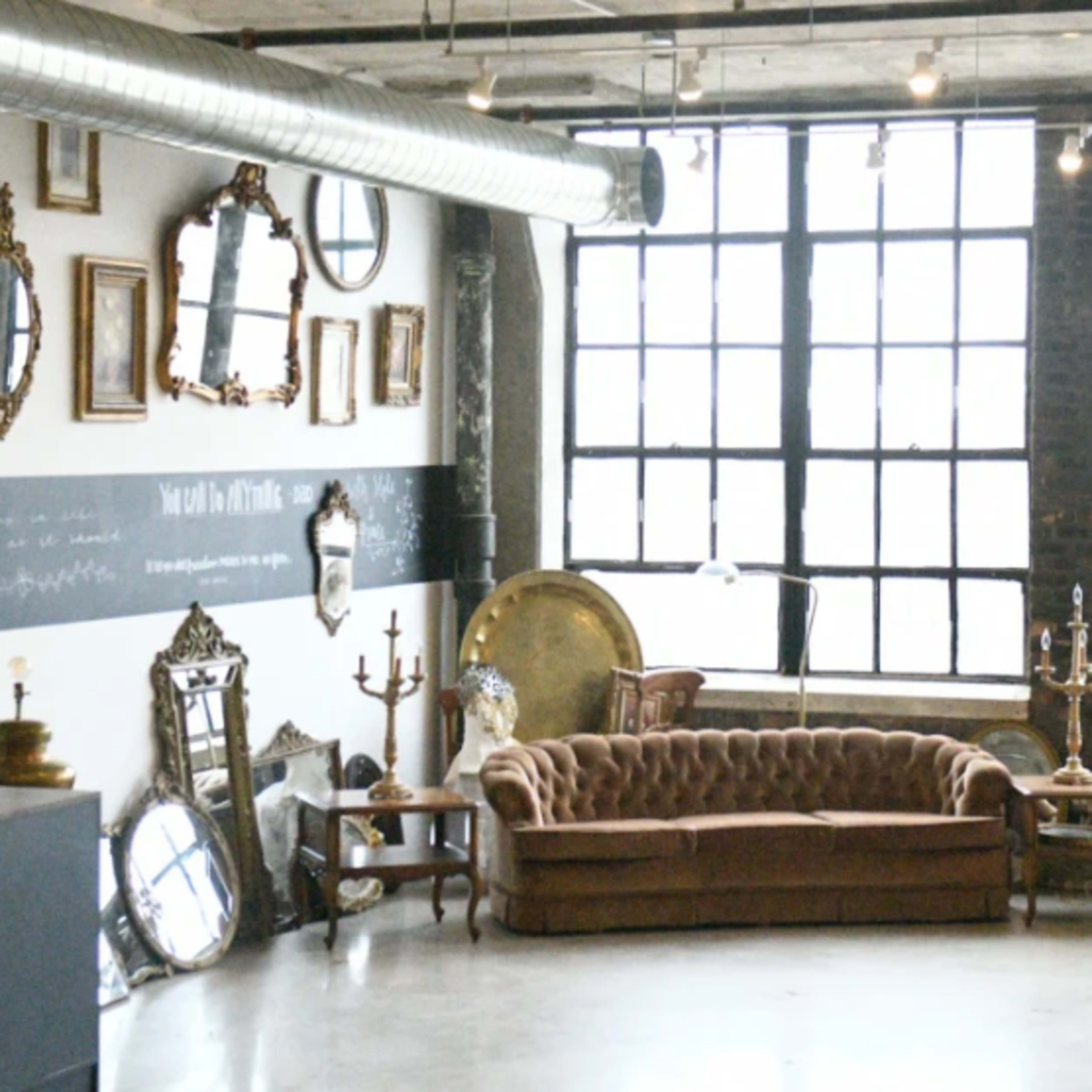 A spacious room with large windows, featuring a mix of vintage furniture and decorative mirrors along the walls.