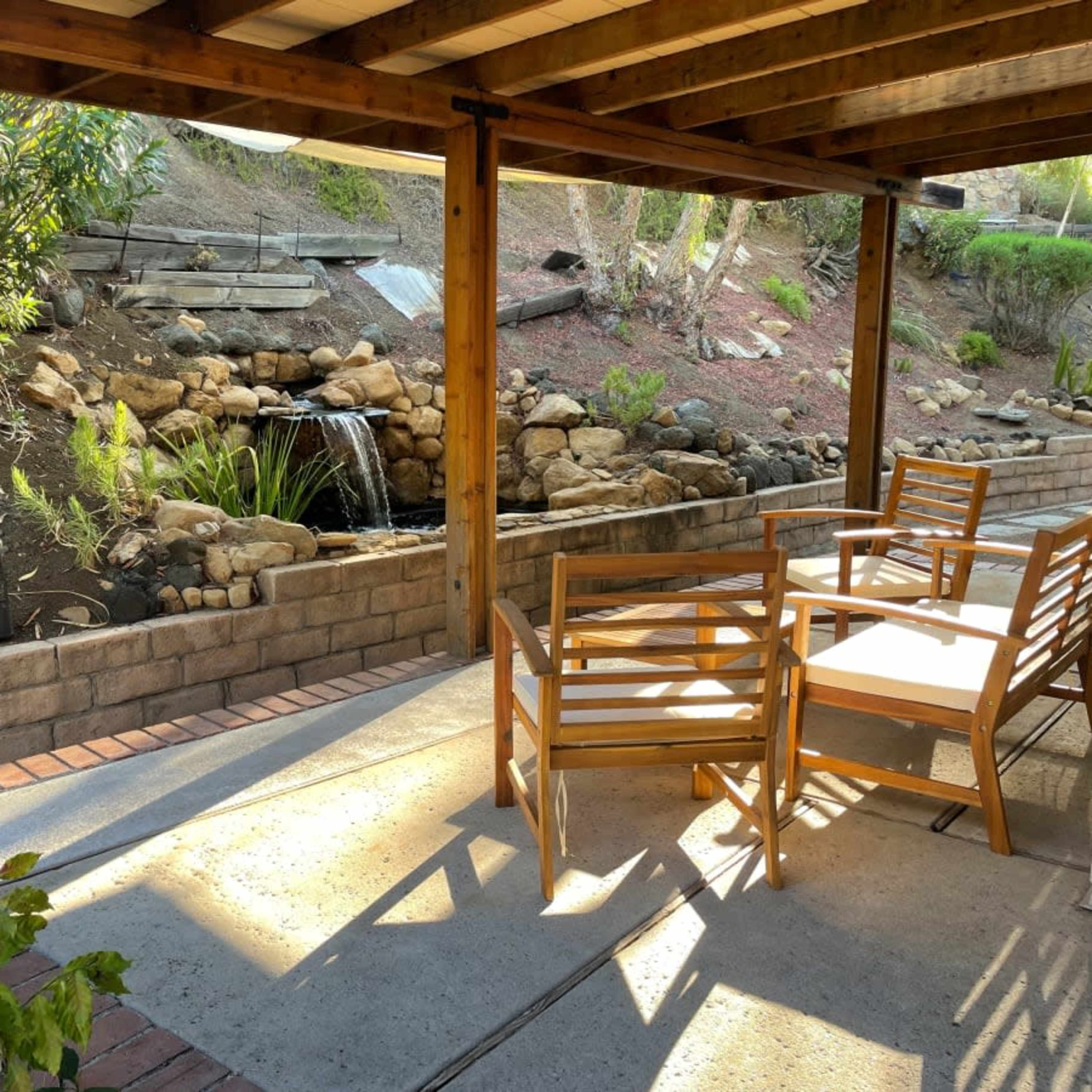 A patio area with wooden furniture overlooking a landscaped garden featuring a small waterfall and rocky elements.