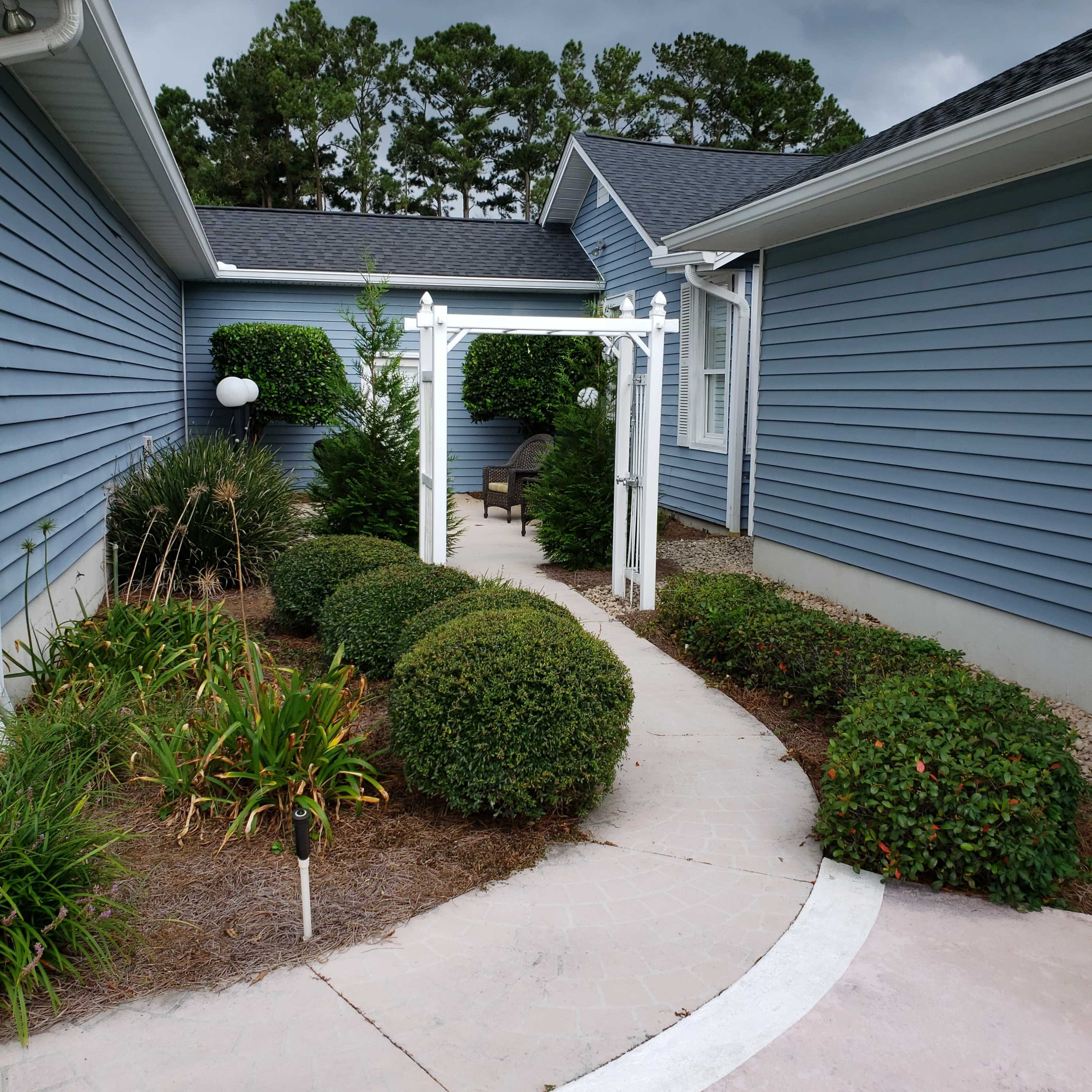 A curved pathway lined with neatly trimmed shrubs leads to a white arbor in a residential garden.