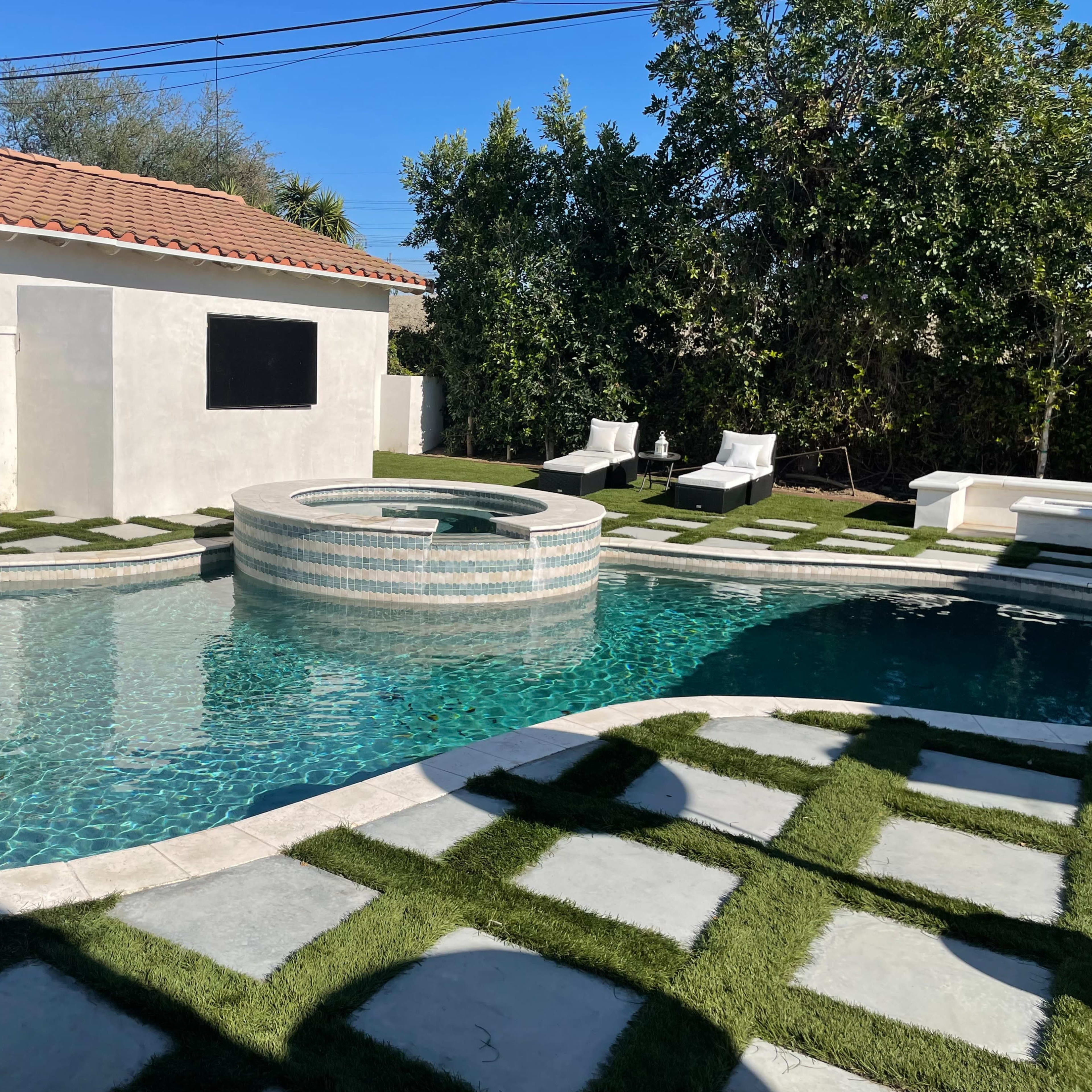 A landscaped backyard featuring a swimming pool with a spa, surrounded by artificial grass and stone pathways, along with lounge chairs positioned nearby.