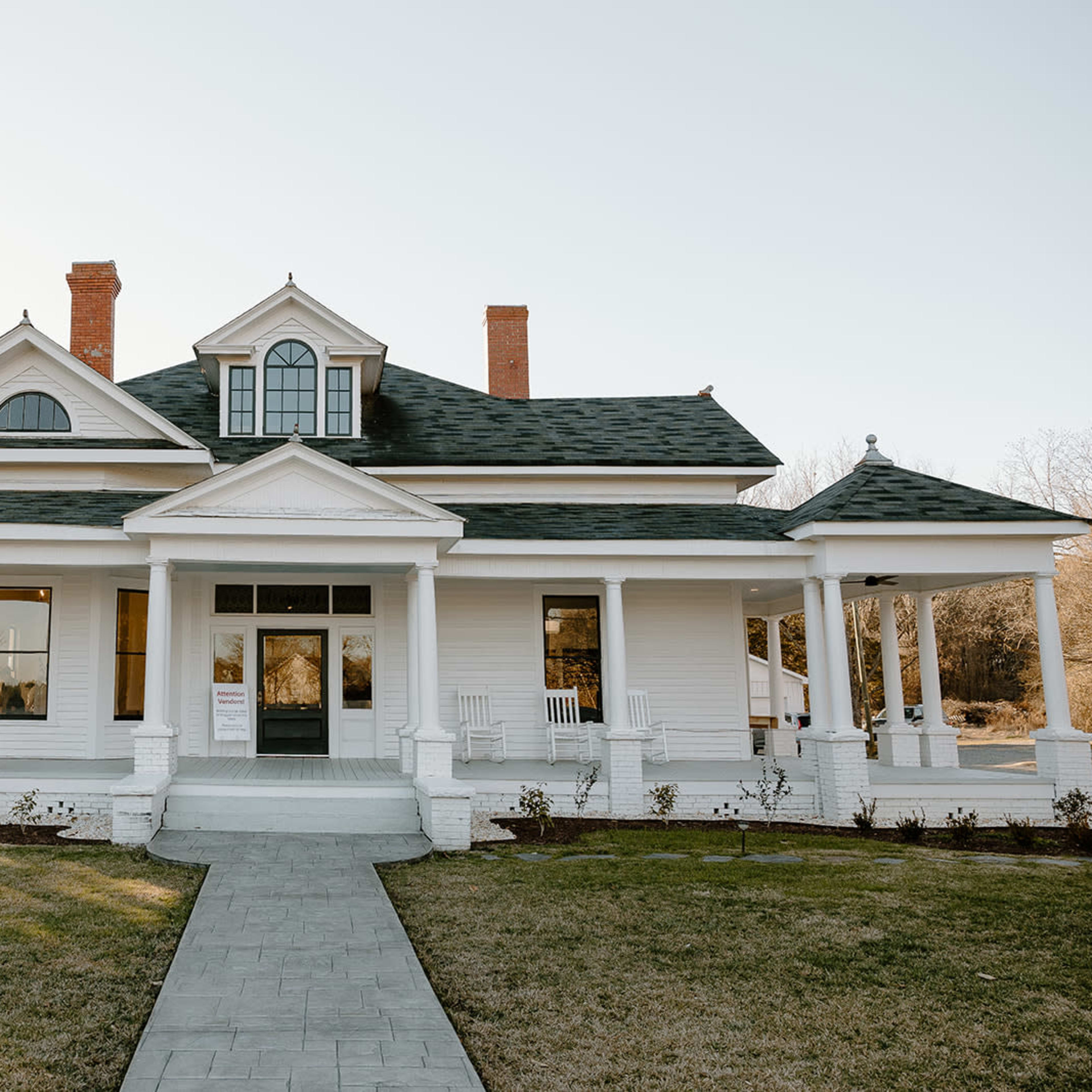 A large white house with a green roof features a central entrance flanked by two porches and surrounded by a well-manicured lawn.