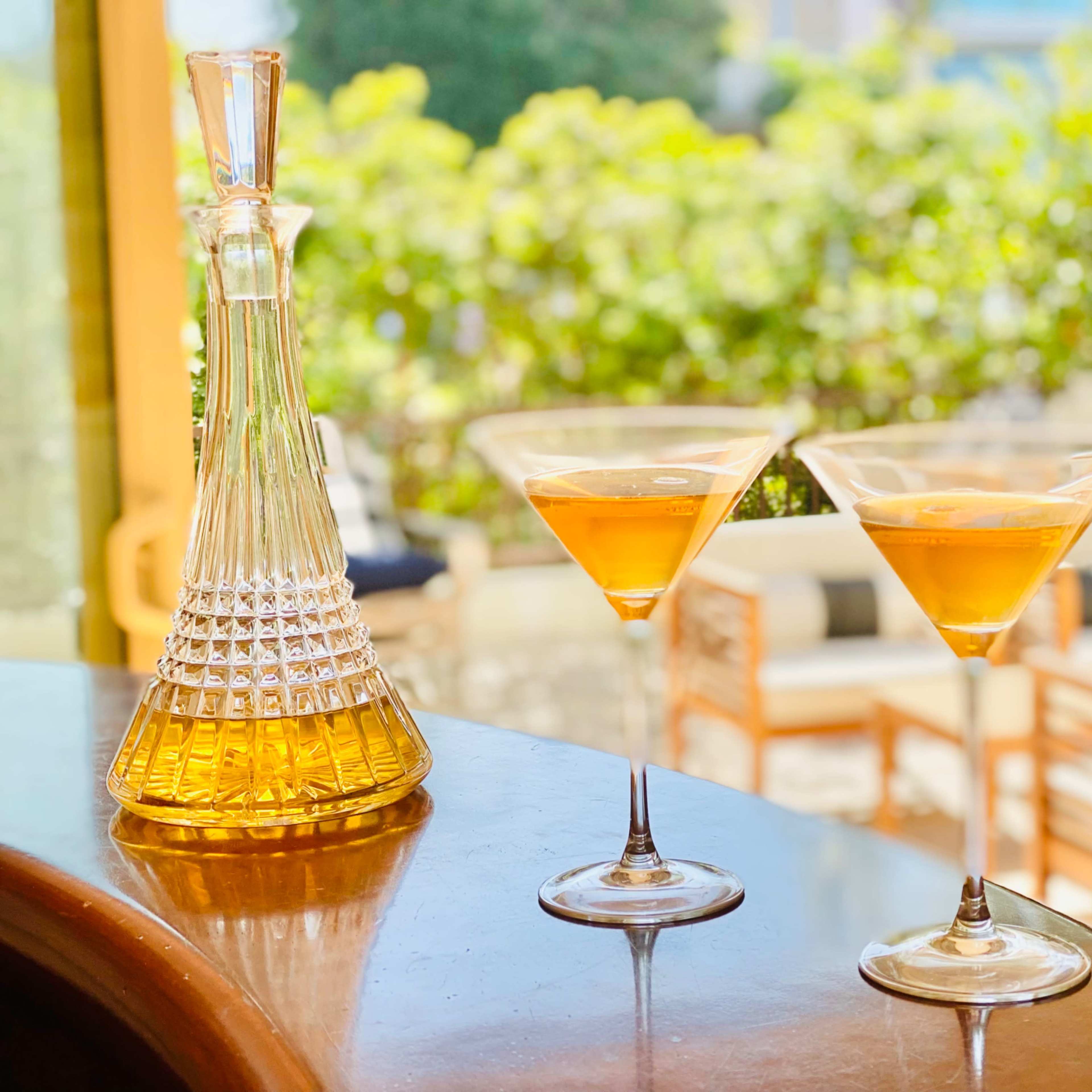 Two martini glasses filled with a golden liquid sit on a bar counter beside a crystal decanter, with a bright outdoor setting visible in the background.