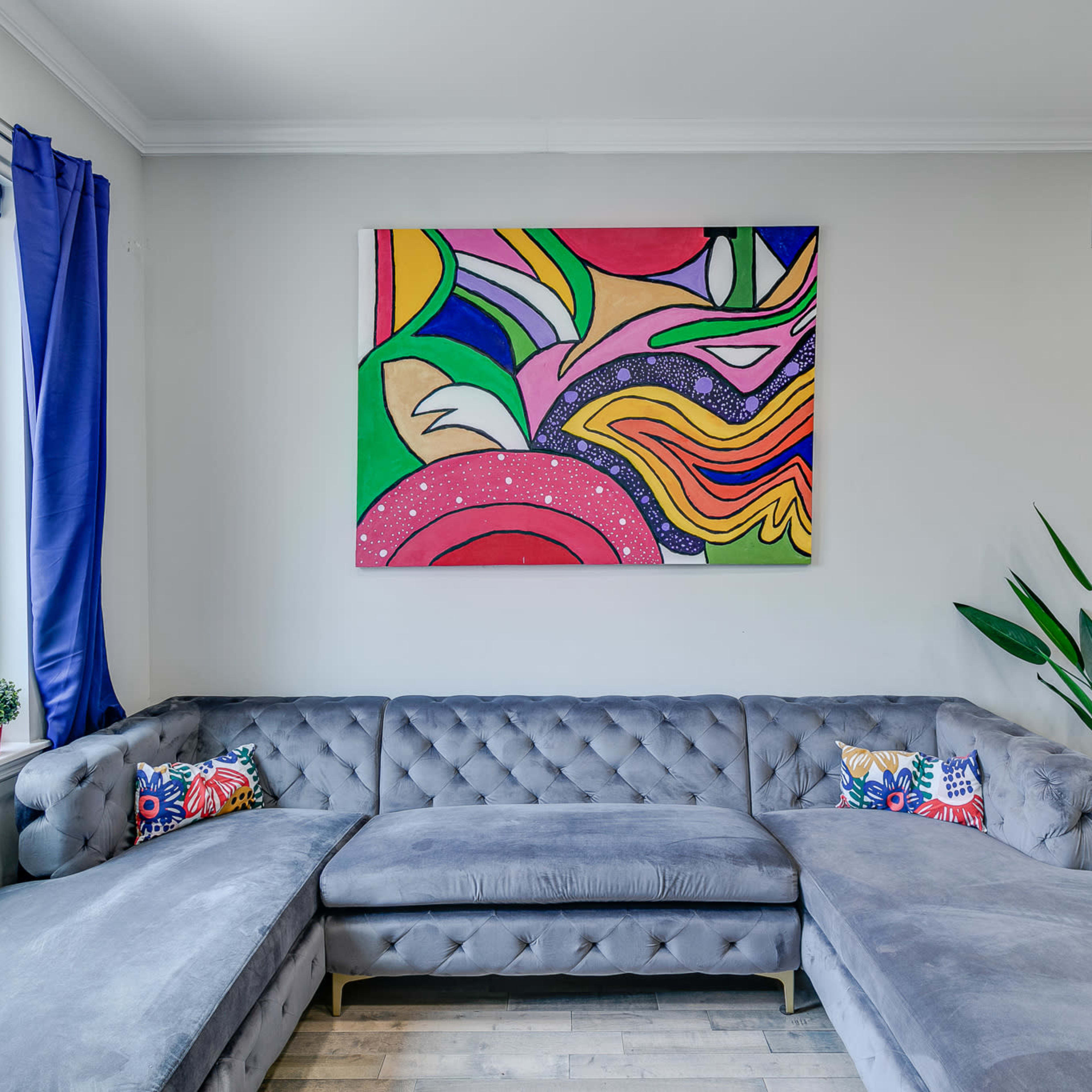 A modern living room features a gray sectional sofa, colorful artwork on the wall, and potted plants by the window.