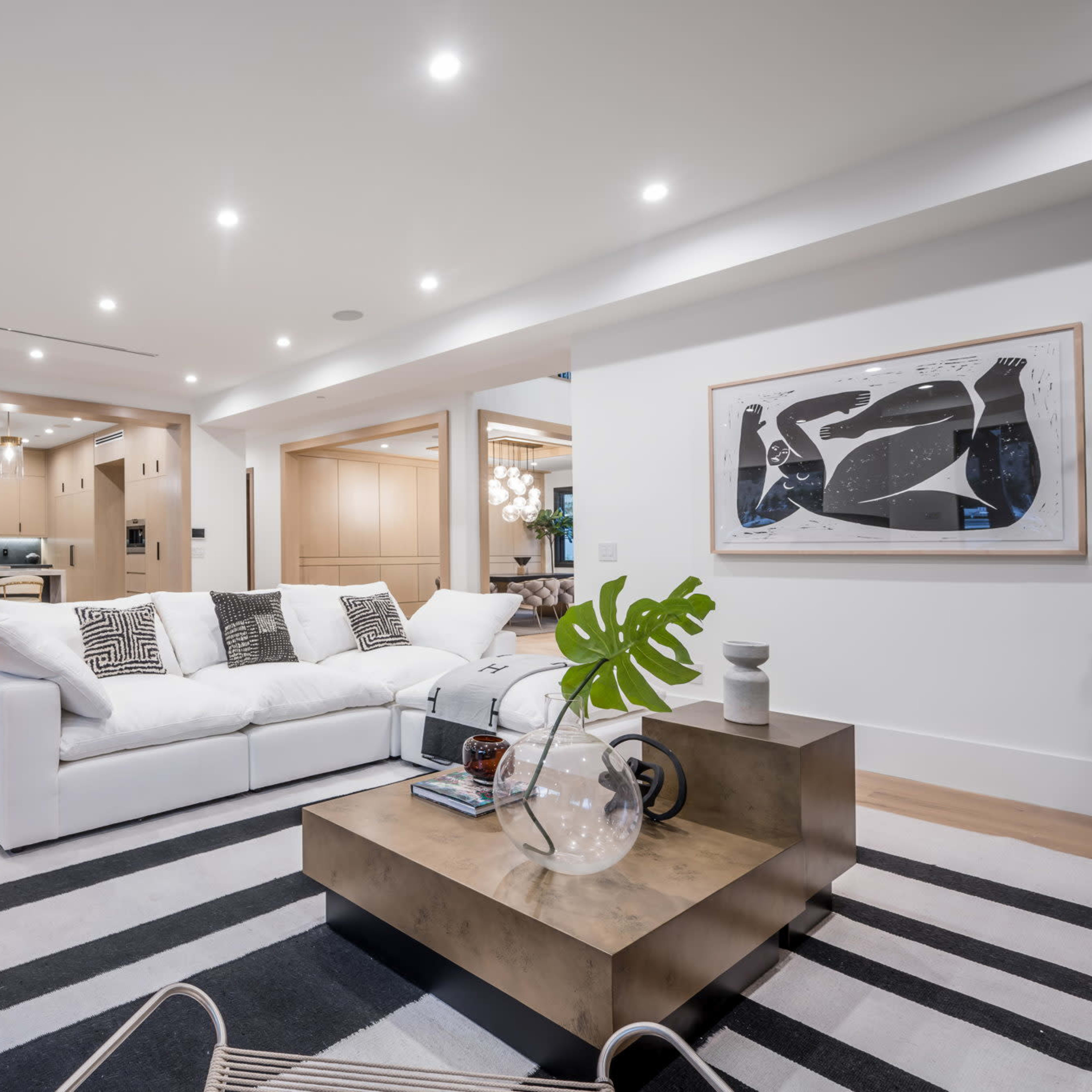 A modern living room featuring a white sofa, a striped area rug, and a coffee table with decorative items, leading into a kitchen with wood cabinetry in the background.
