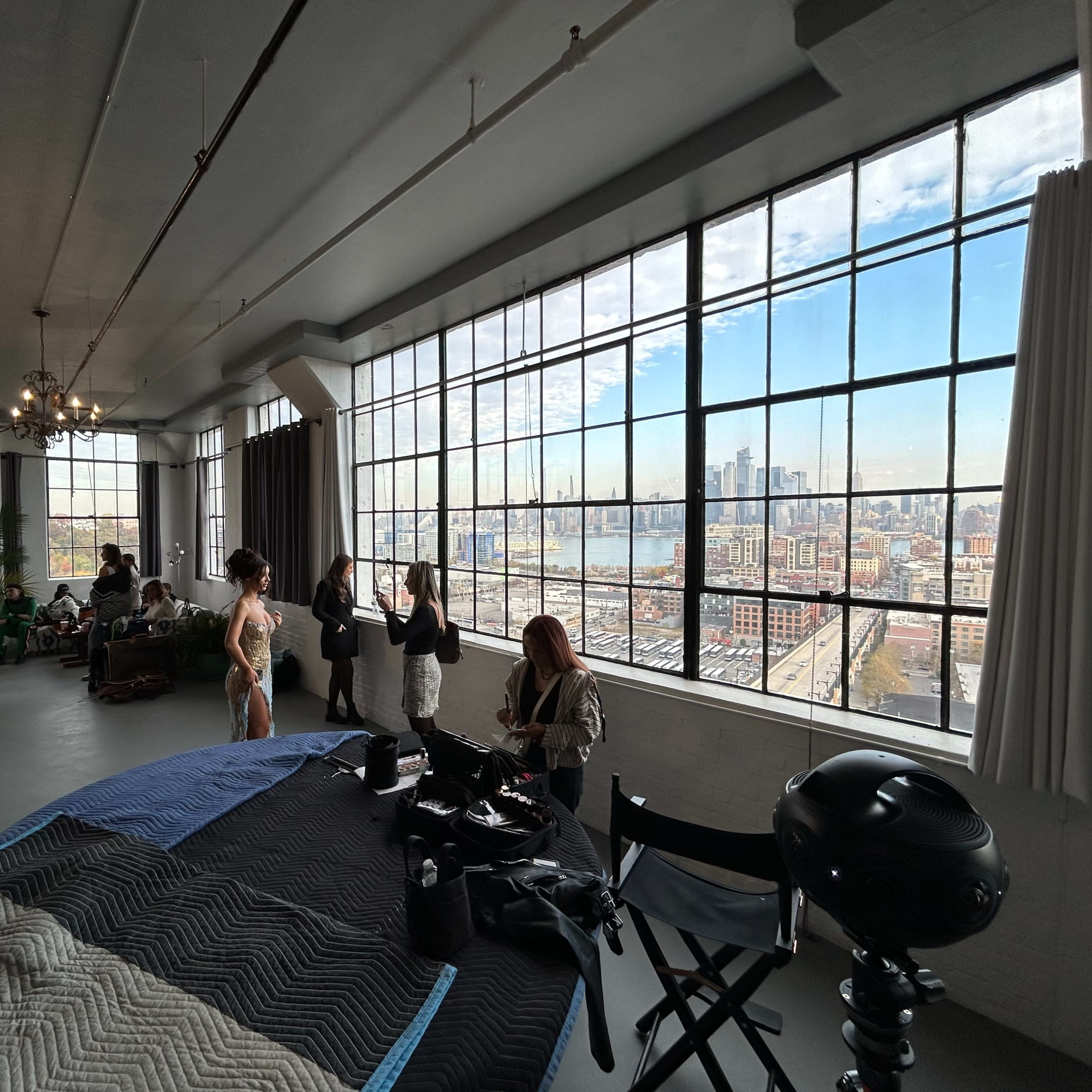 A bright studio with large windows overlooks an urban skyline, while people are preparing and organizing equipment inside.