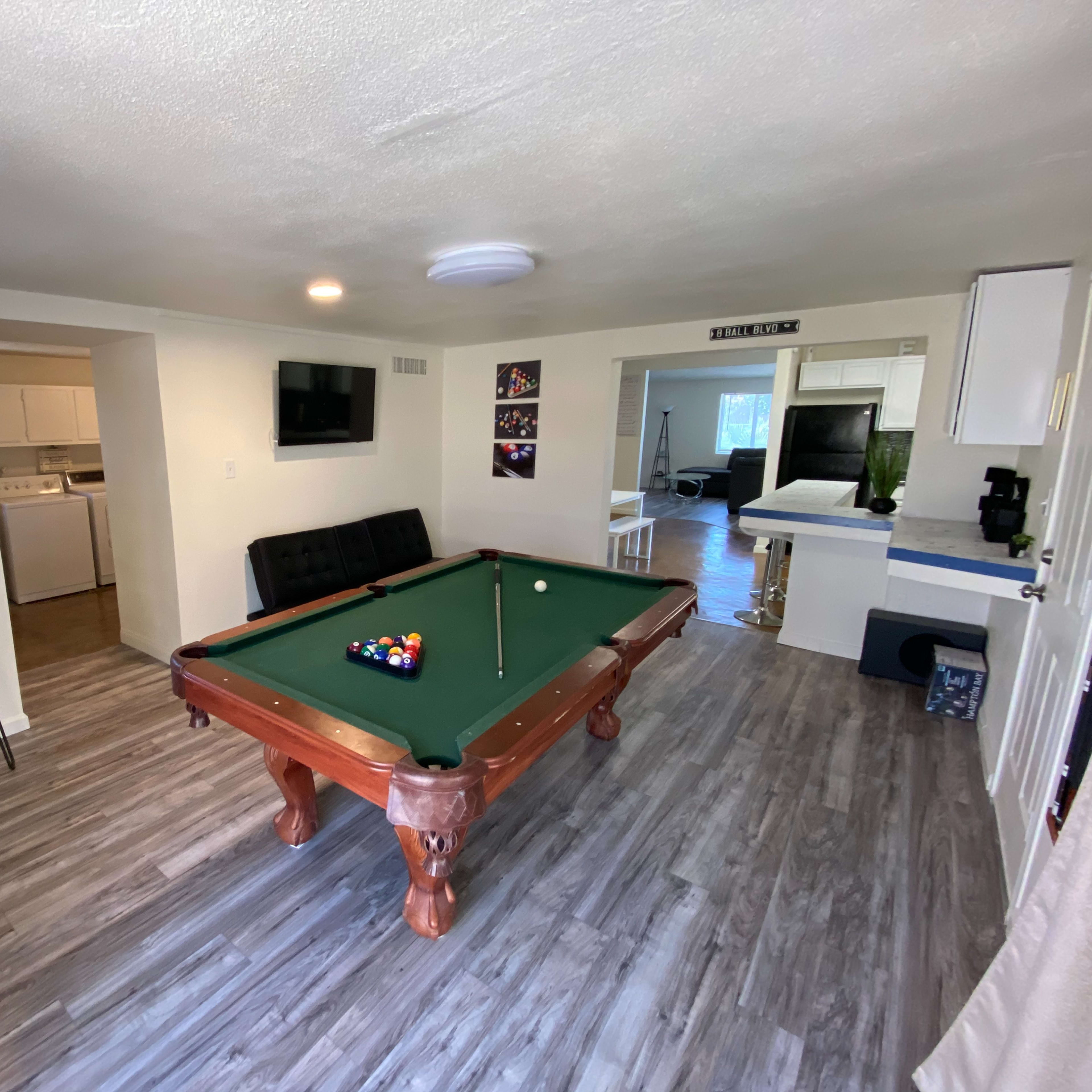 A room featuring a pool table, bar stools, and a kitchenette visible through an open doorway.