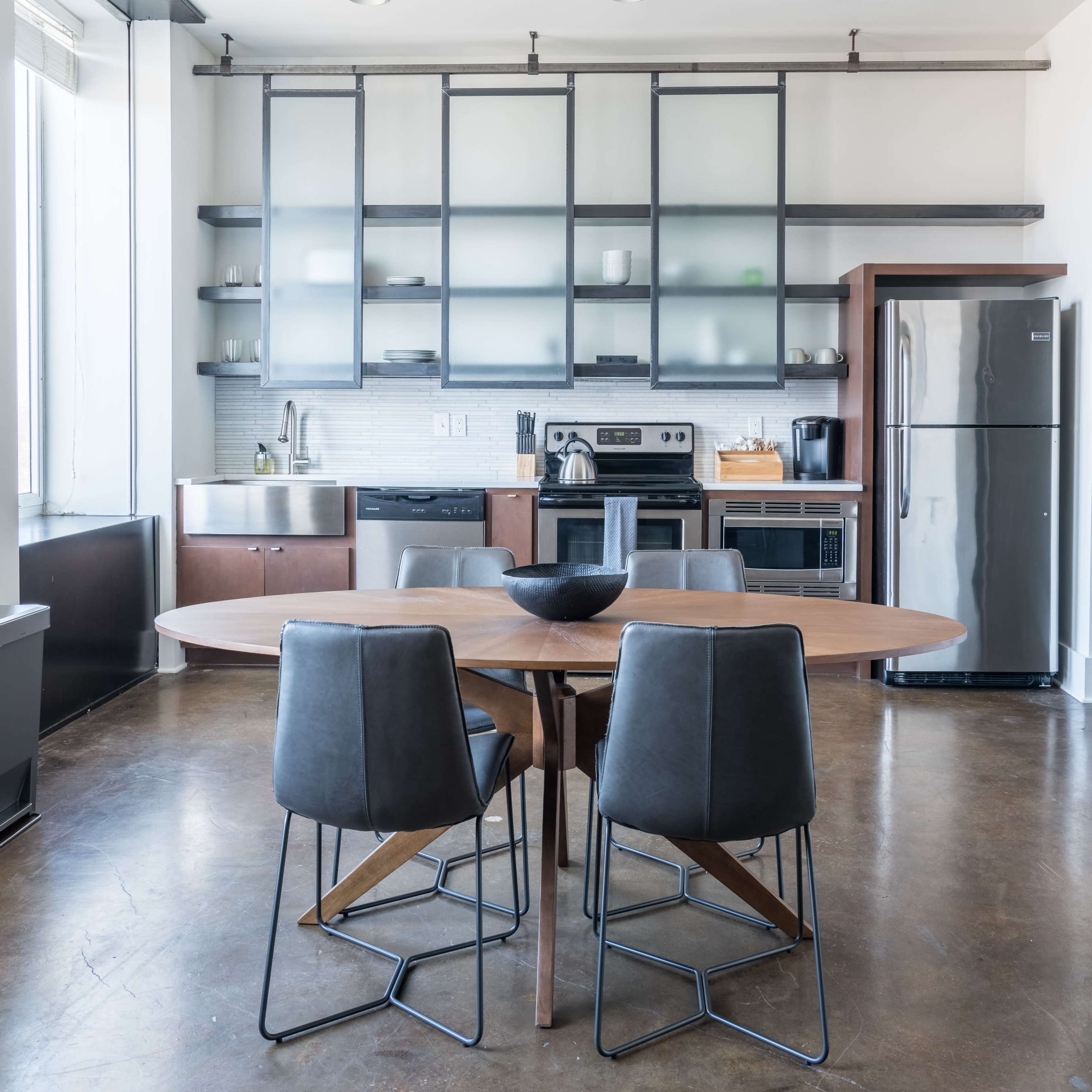 A modern kitchen with a large round wooden table and four black metal chairs, along with stainless steel appliances and glass-front cabinetry.