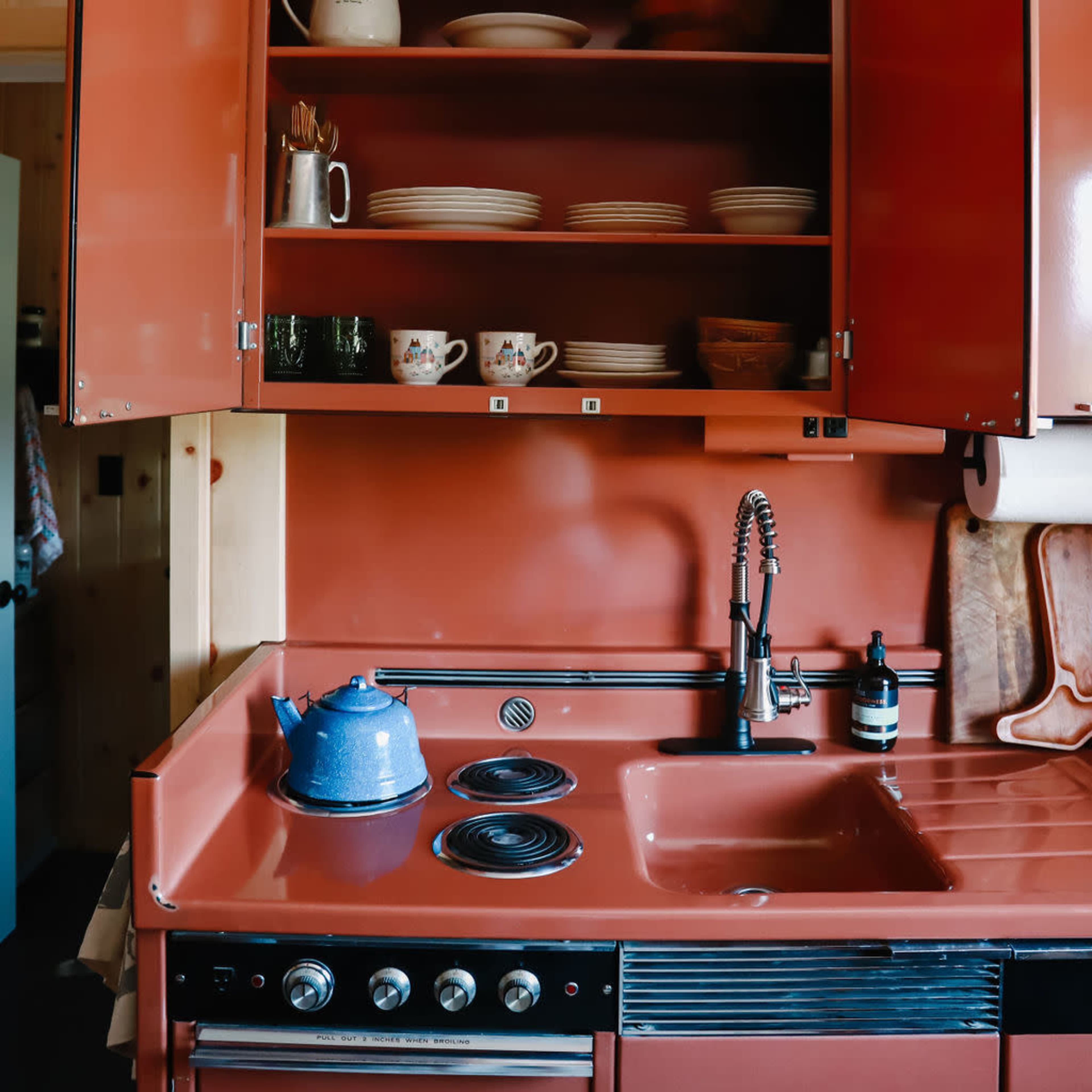 A vintage kitchen with red cabinets, a blue kettle on the stove, and dishes neatly arranged on open shelves.