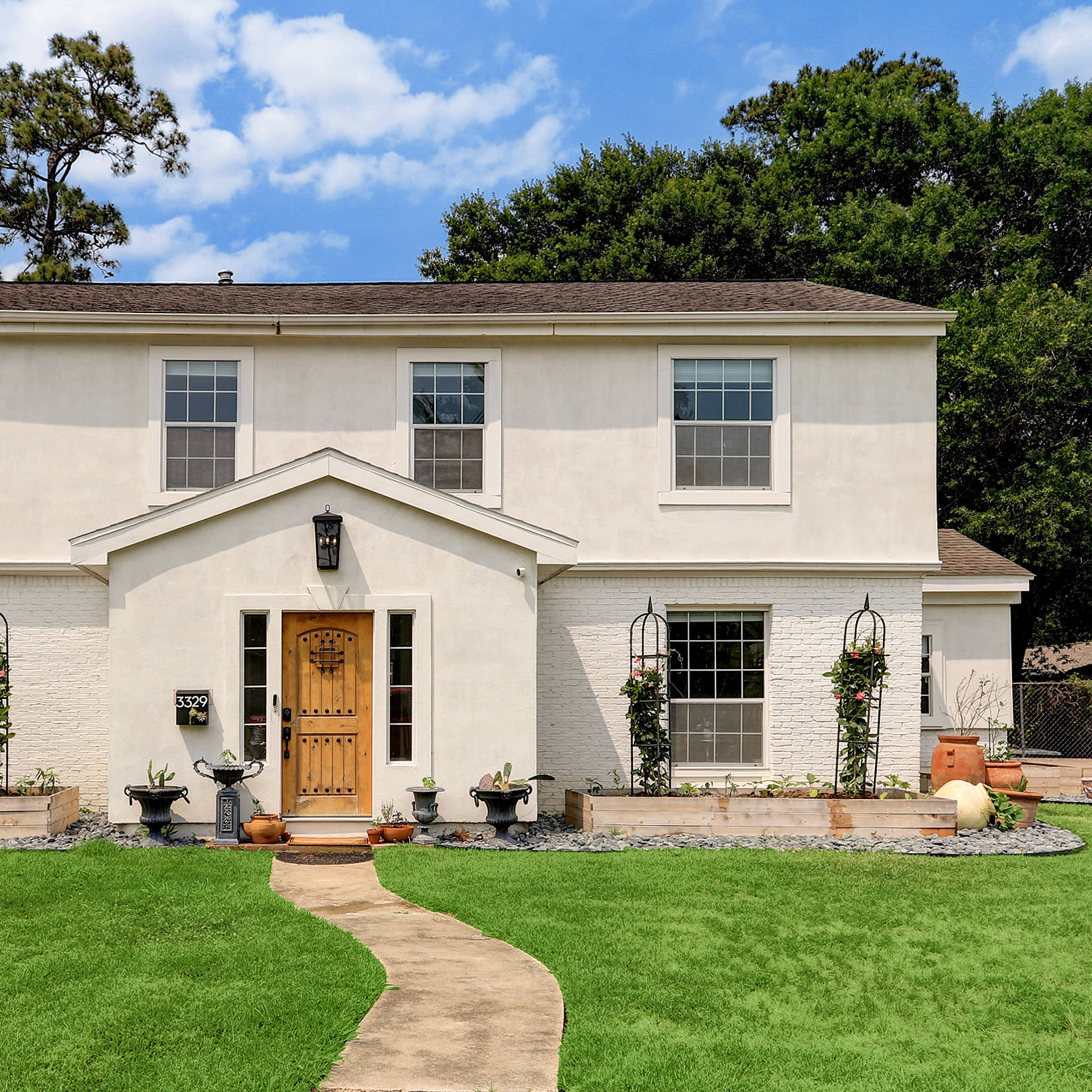A two-story white house with a central entrance and surrounding lush lawn features potted plants and decorative elements near the front path.