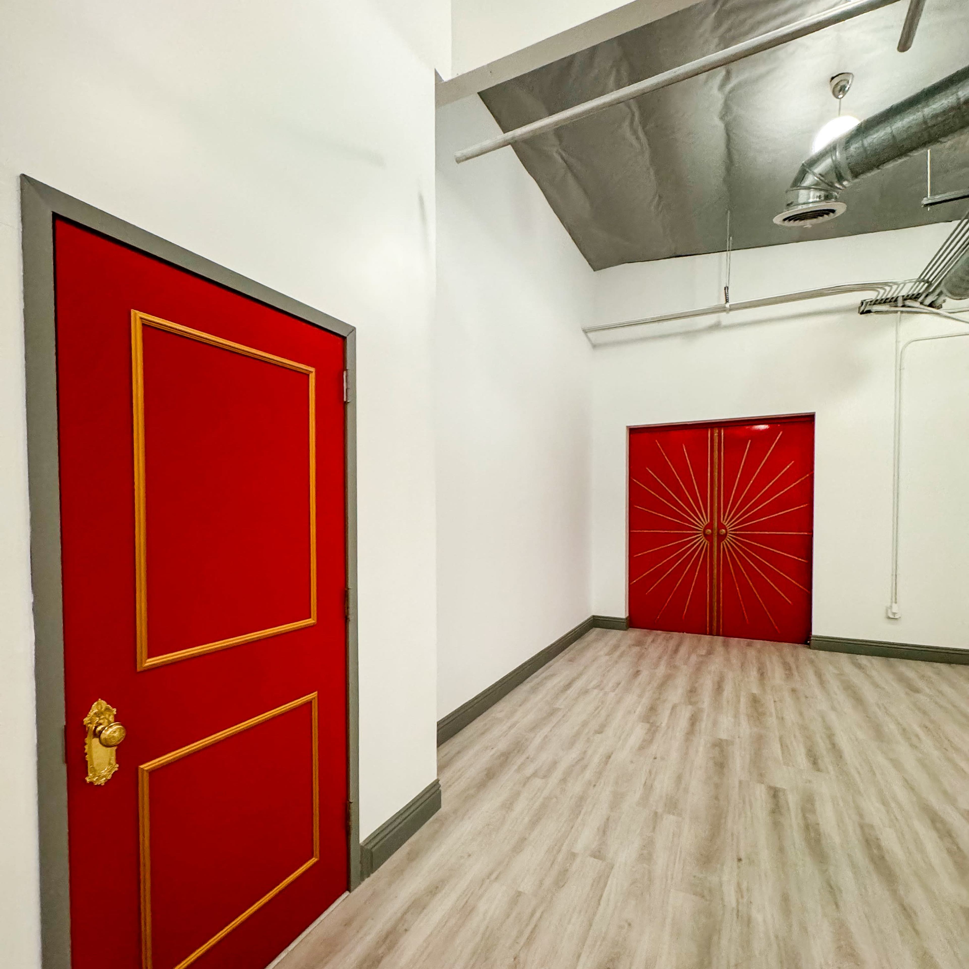 A hallway with two red doors, one featuring a simple frame and the other adorned with a sunburst design, against white walls and light wooden flooring.