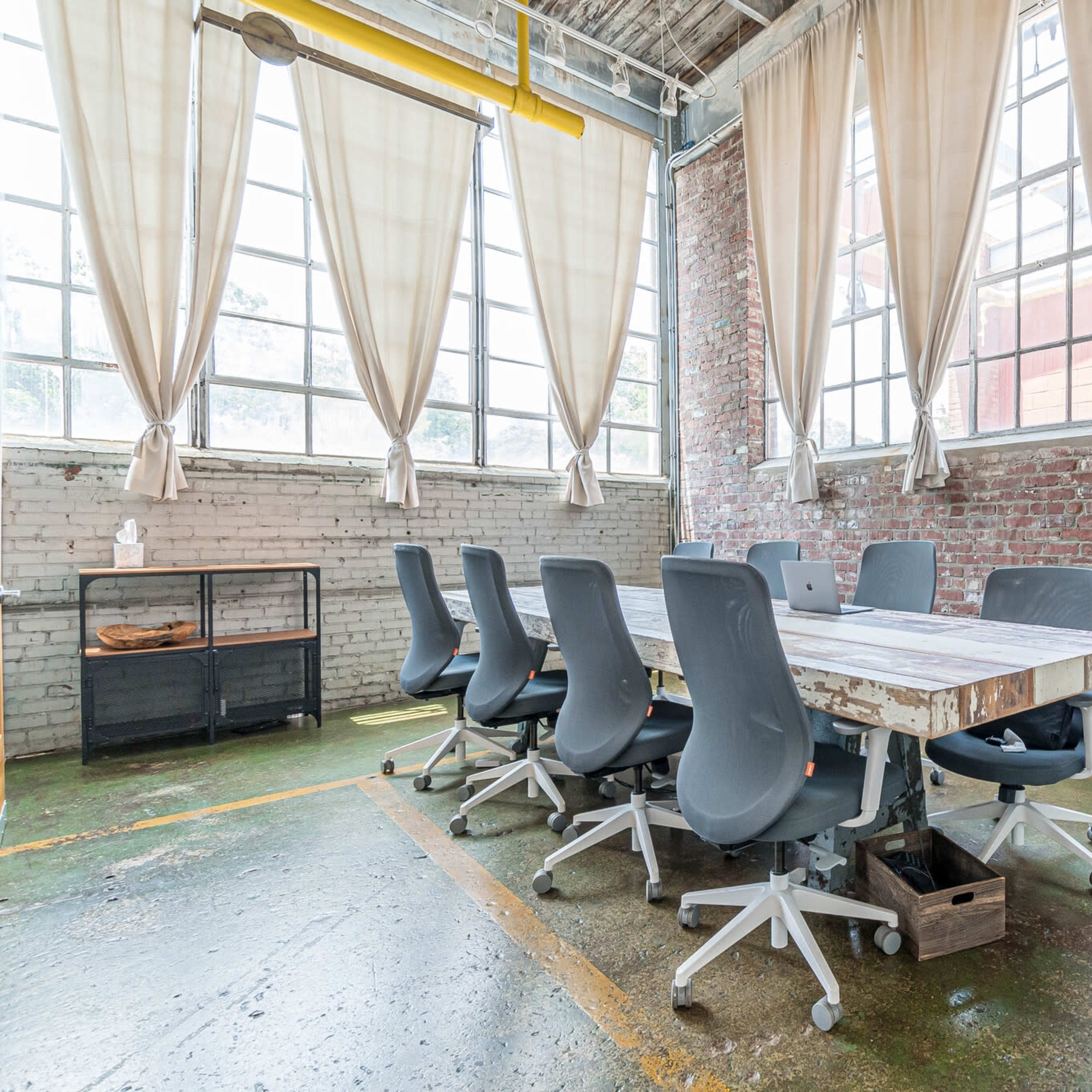 A long wooden table surrounded by gray chairs is set in a room with large windows and exposed brick walls.
