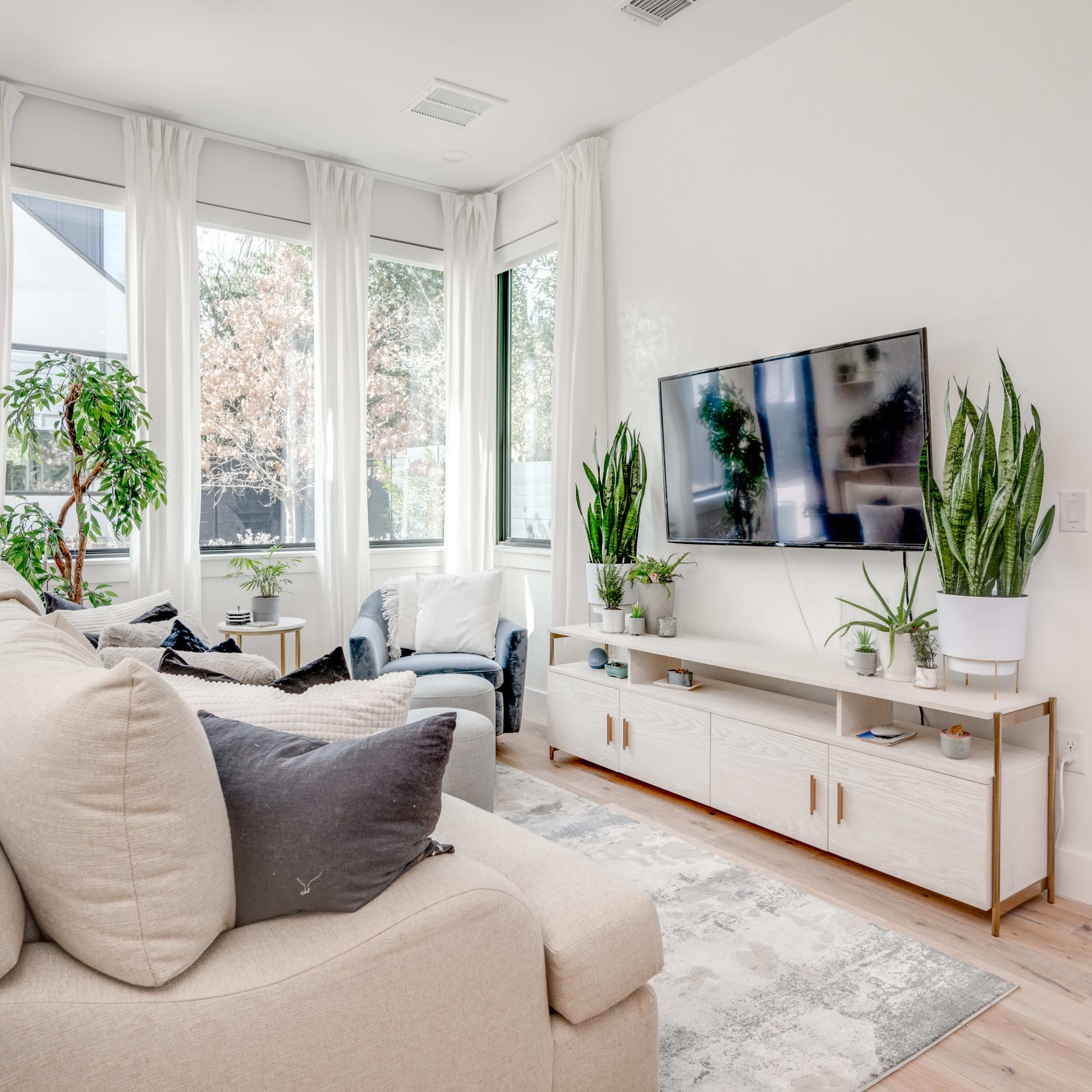 A modern living room features a light-colored sofa, a media console with plants, and large windows allowing natural light.