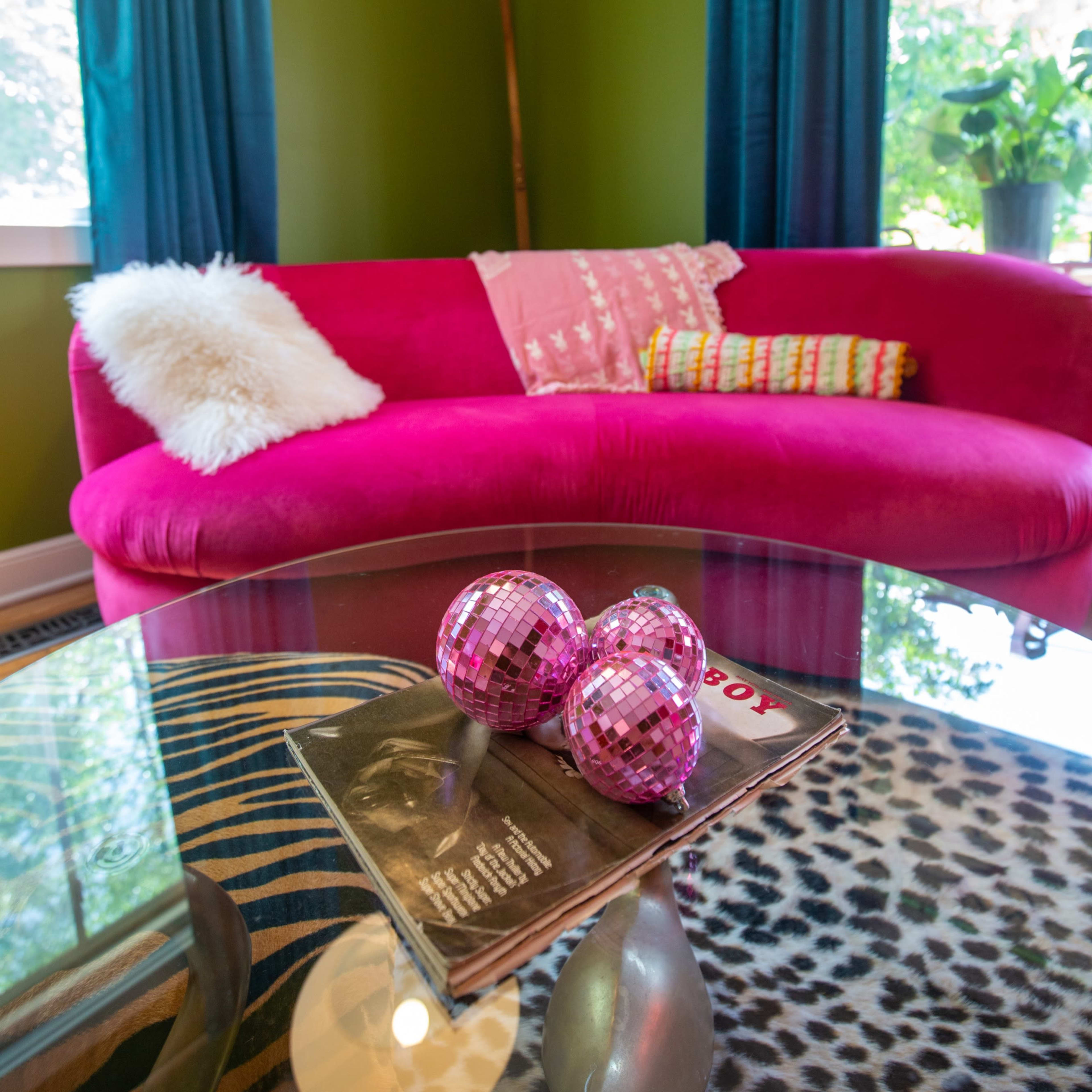 A pink sofa with colorful pillows sits in a room featuring green walls and large windows, with a glass coffee table displaying magazines and decorative disco balls.