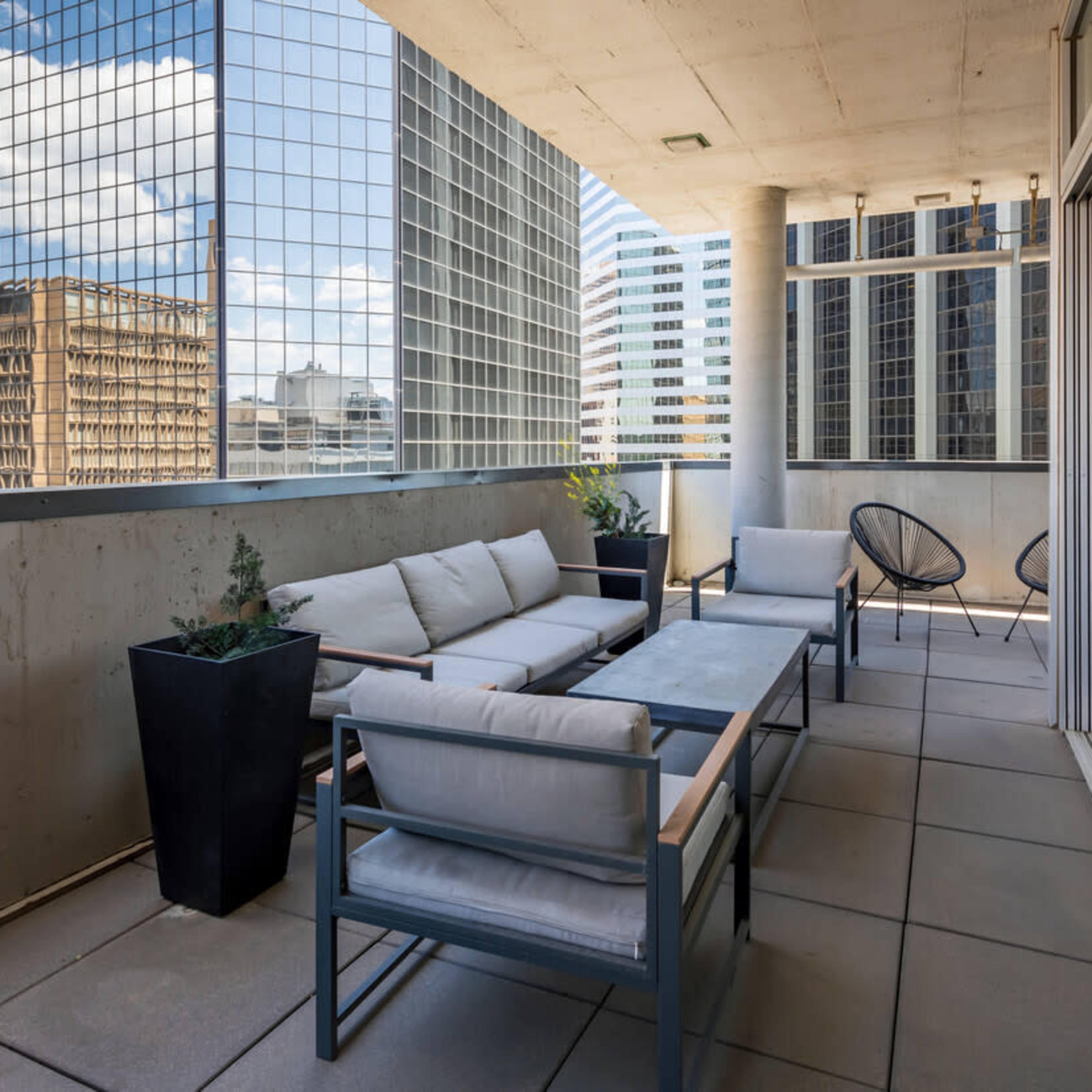 A modern balcony features a gray seating area with cushions, a rectangular table, and black planters, overlooking tall glass buildings.