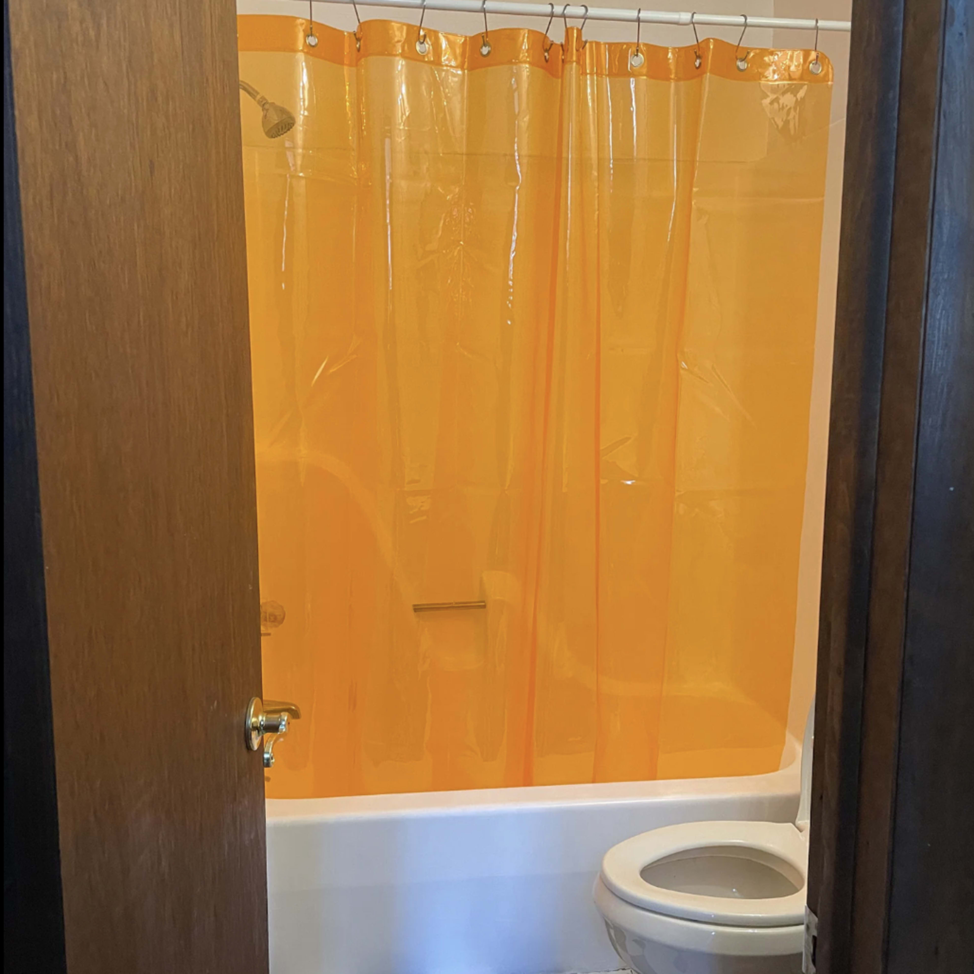 A bathroom features a toilet and a shower area separated by an orange plastic curtain, with a black and white checkered floor.