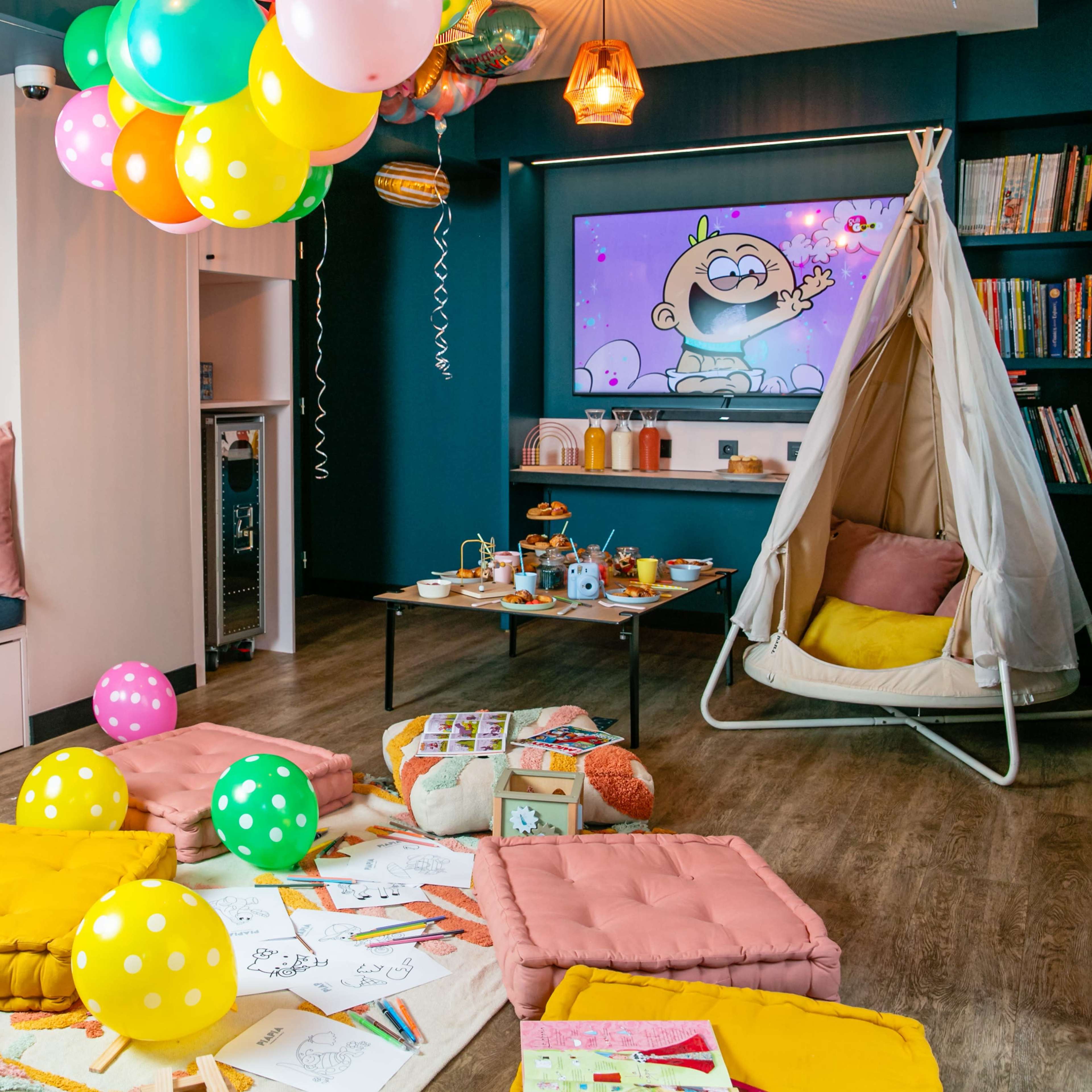 A colorful children's playroom features a swinging chair, various plush cushions on the floor, and a decorated table with snacks and art supplies, all under balloons and a television displaying animated content.