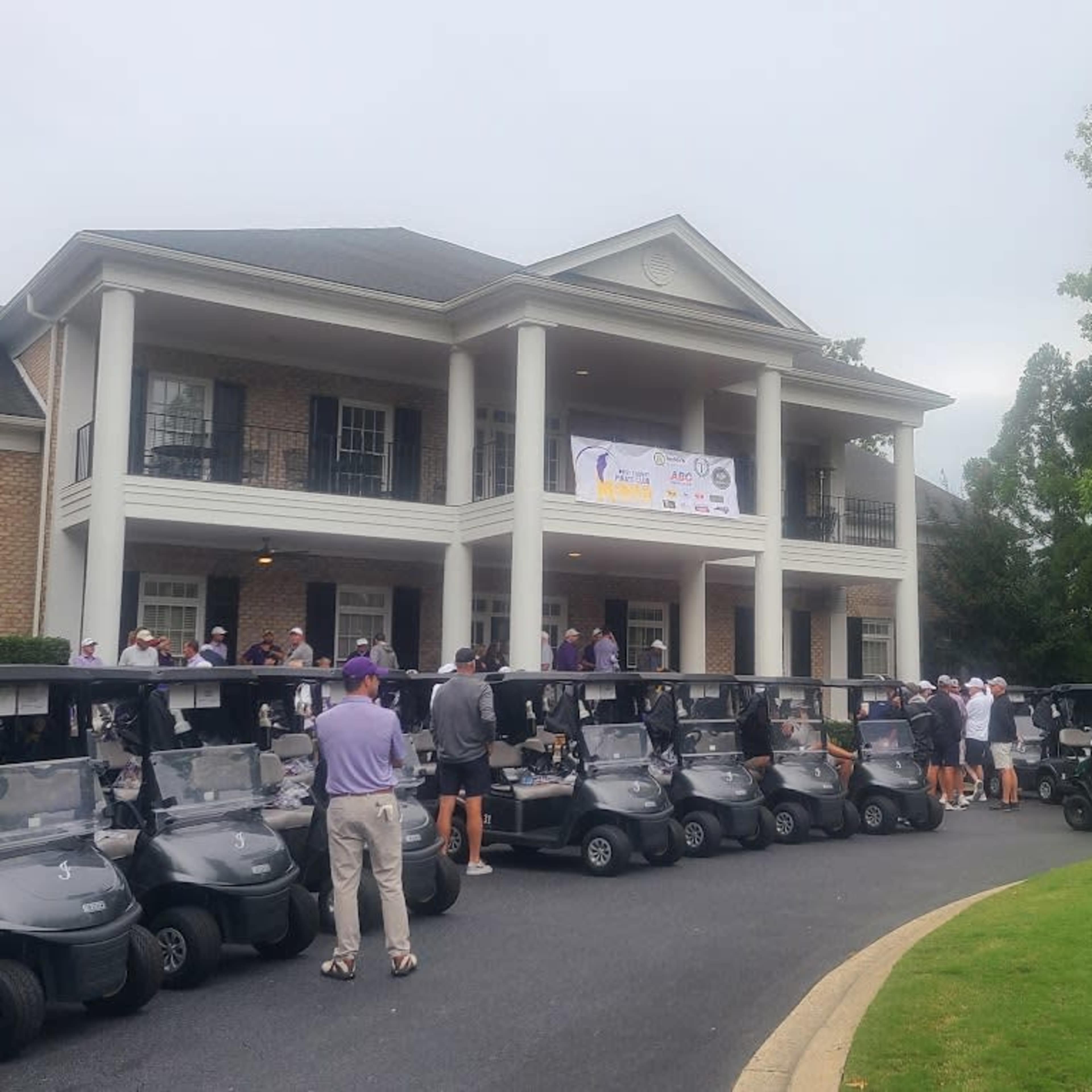 A row of golf carts is lined up in front of a brick building with a porch, where people are gathered for an event.