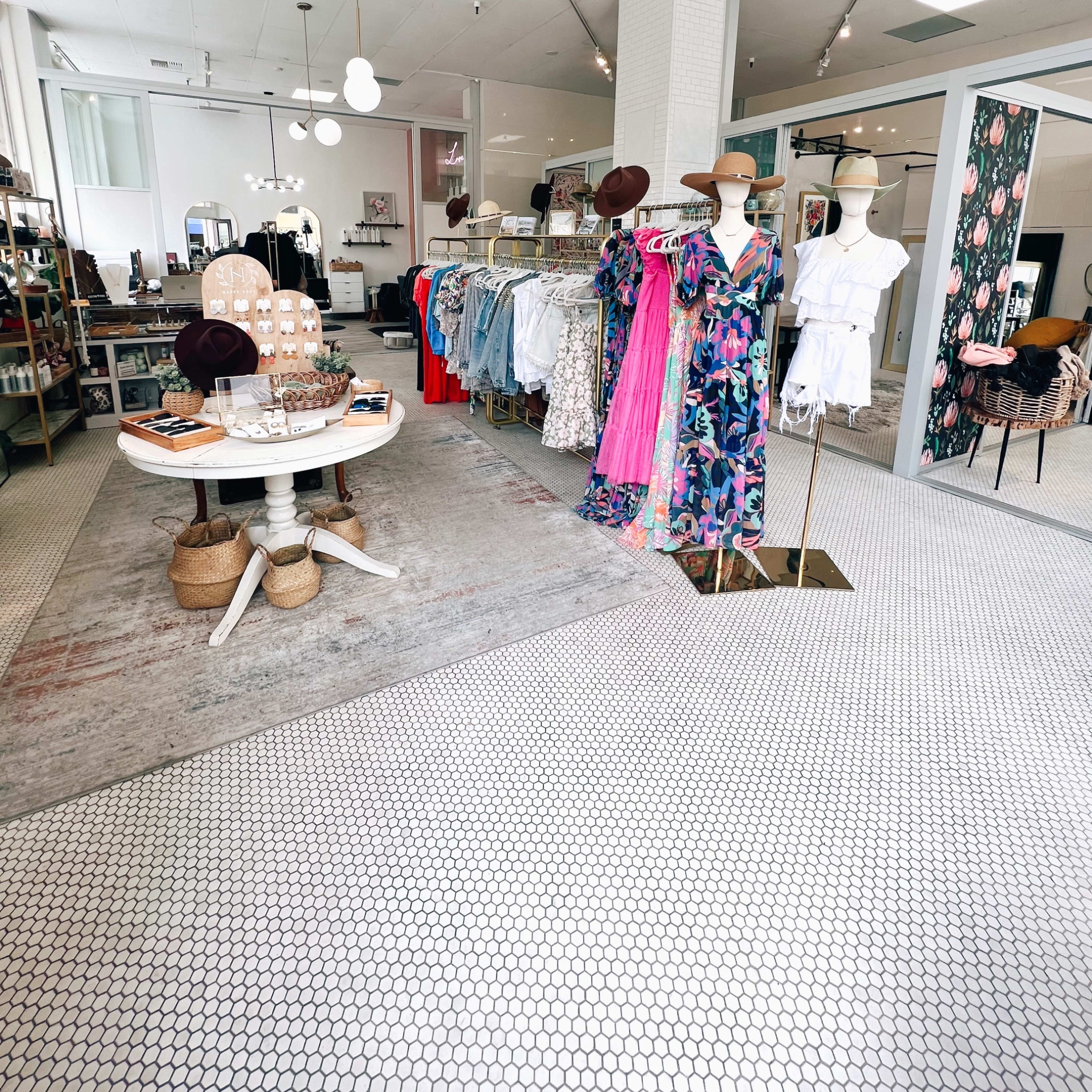 The interior of a boutique featuring clothing racks, a round table, and decorative elements, with a light-colored tile floor and bright lighting.