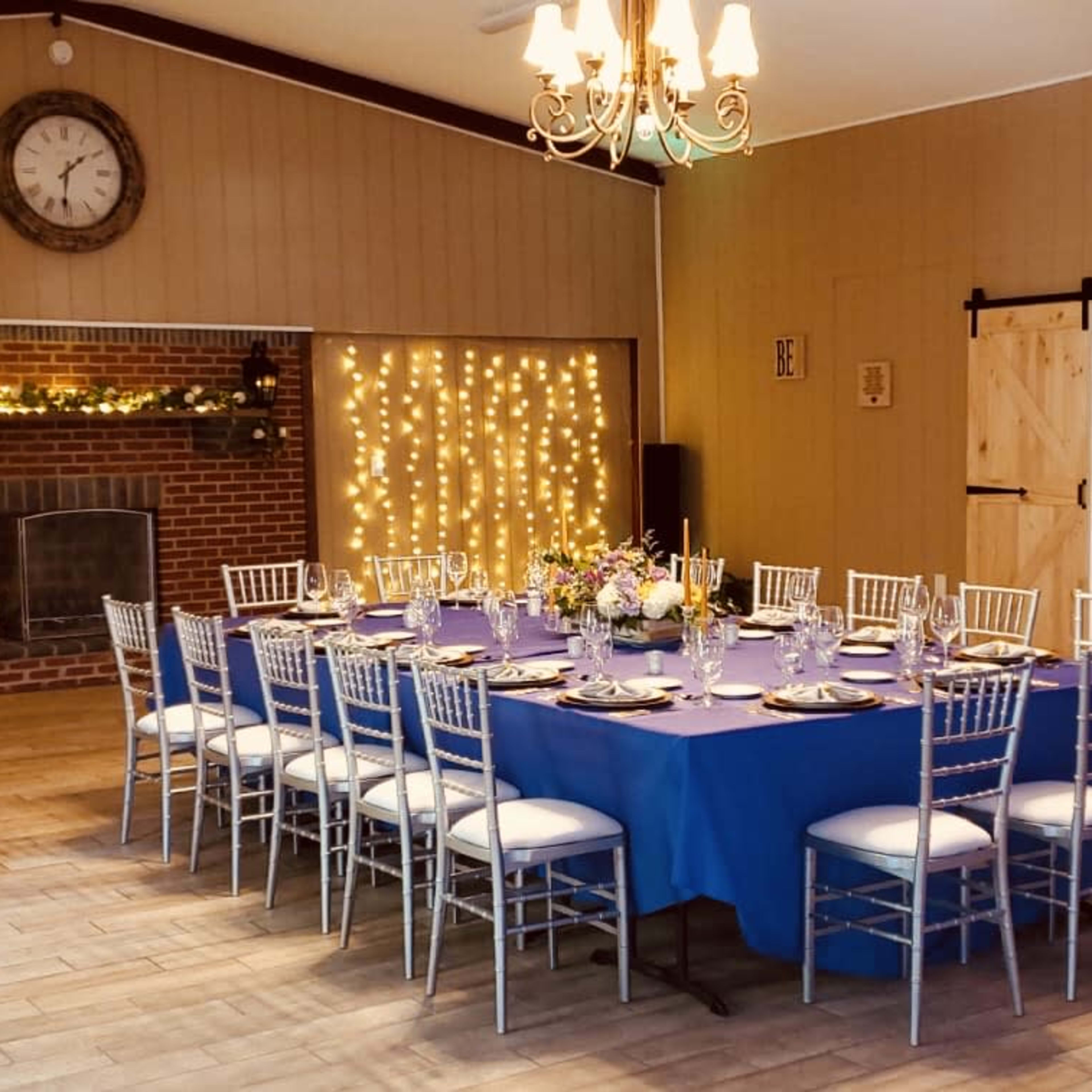 A dining area features a round table set with floral centerpieces and surrounded by silver chairs, with a brick fireplace and string lights in the background.