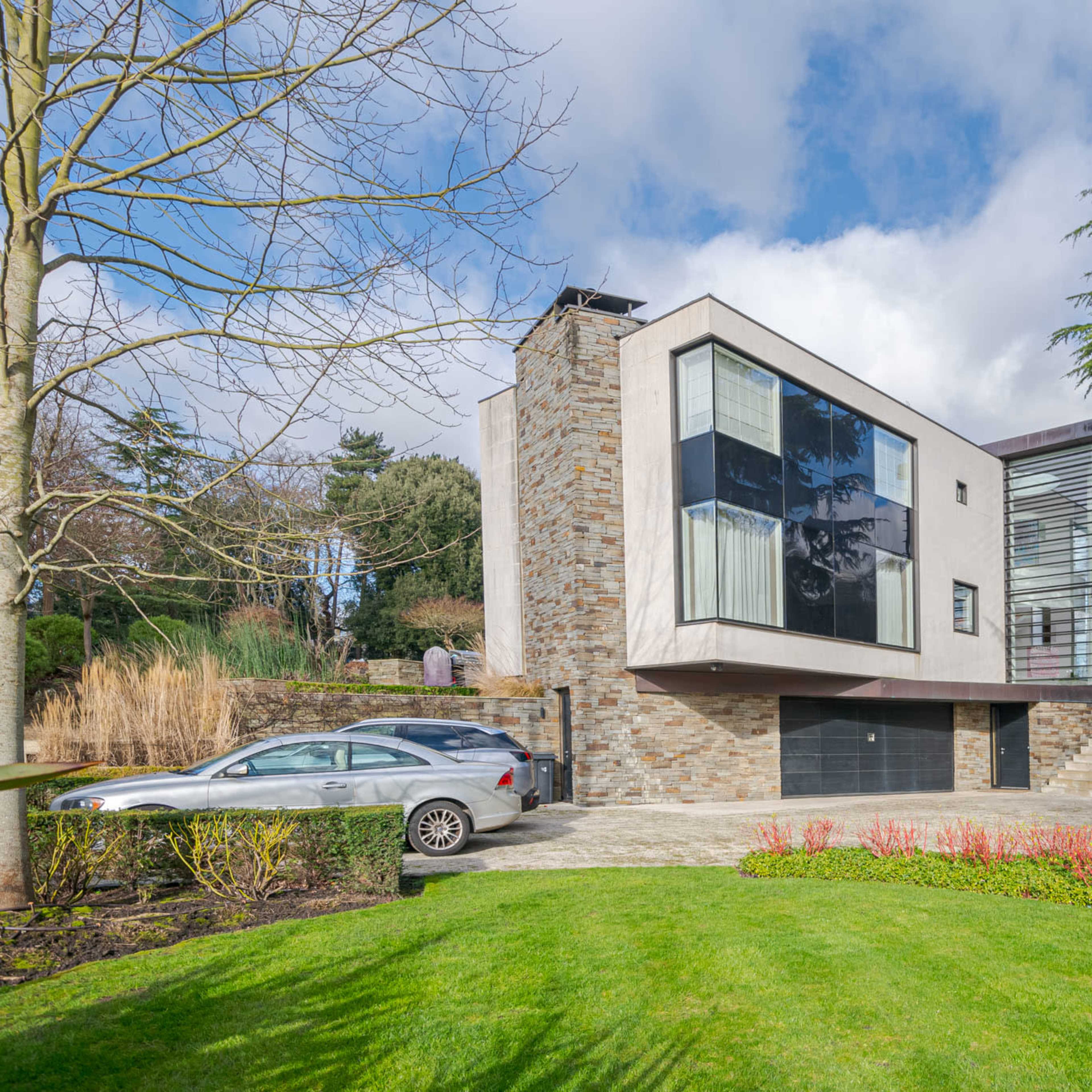 A modern, two-story house featuring large windows and a stone facade is set amidst landscaped greenery and a driveway.